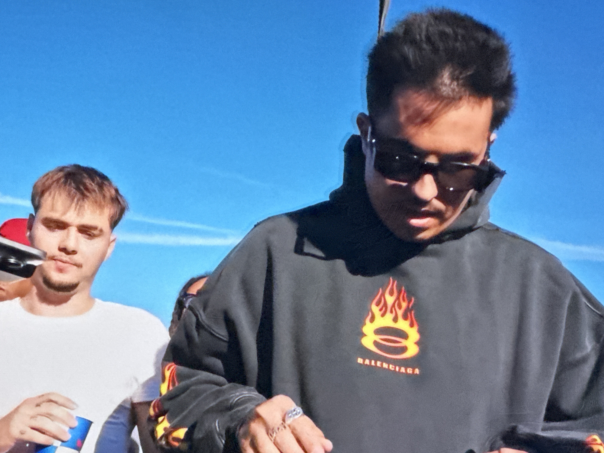 The image shows two men outdoors under a clear blue sky. The man in the foreground is wearing a dark grey Balenciaga hoodie with a flame design and sunglasses. He has short dark hair and is wearing rings on his fingers. The second man is standing slightly behind and to the left, wearing a white t-shirt and holding a can of Red Bull. The location in Bragança, Portugal is unknown and not visible.