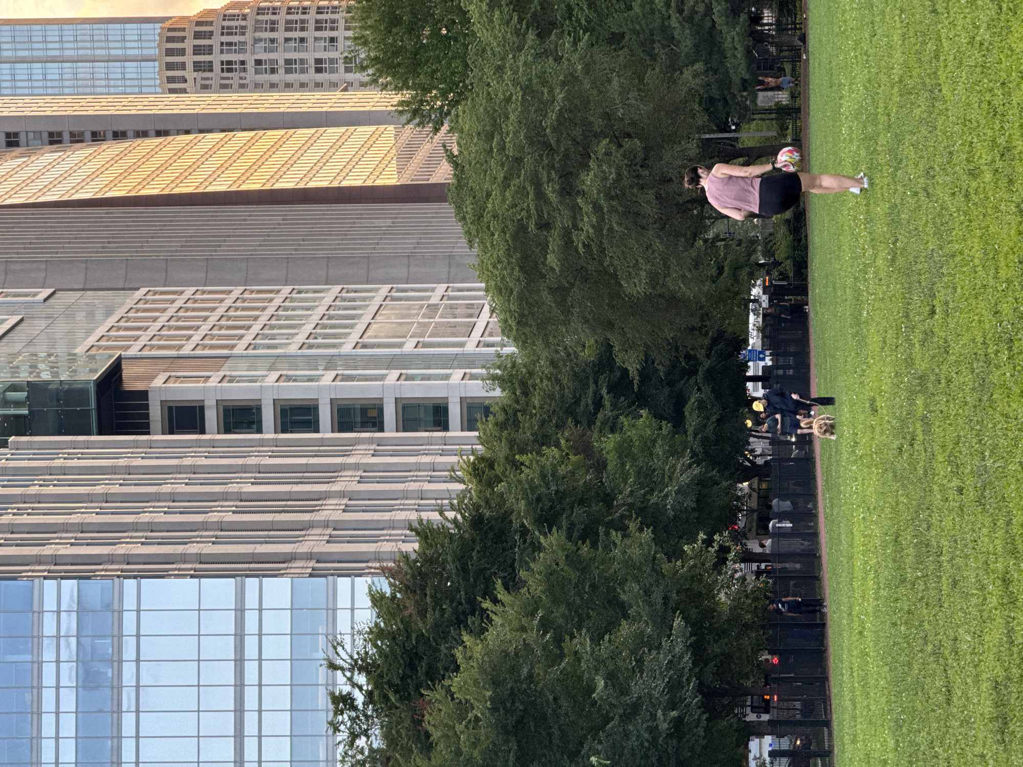 In a city park in Chicago, Illinois, a woman wearing a purple tank top and black shorts is standing on a green grassy field, holding a colorful soccer ball. Behind her, a dense line of trees separates the park from a row of tall, modern skyscrapers. In the background, several people are walking and standing near a fence, and a golden retriever on a leash is being walked by a person wearing a yellow hard hat. The sun appears to be setting, casting a warm glow on the windows of the buildings.