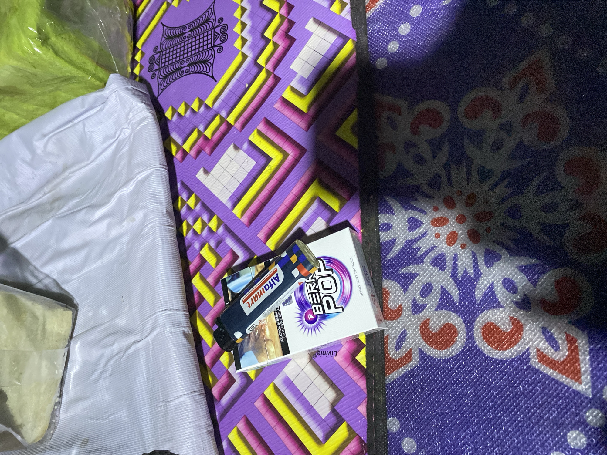 In Lamtui, Indonesia, a pack of "BERRY POP" cigarettes with a blue and white design, featuring a burst-like graphic and the text "BERRY MINT CAPSULE" is placed on a purple mat with a geometric pattern of yellow, pink, and white squares. A blue lighter with the "Alfamat" logo is resting on top of the cigarette pack. The scene is illuminated by a single light source, casting shadows. In the background, two large white and green plastic-wrapped objects are visible, one appearing to contain a light yellow substance. The mat itself has a woven texture.