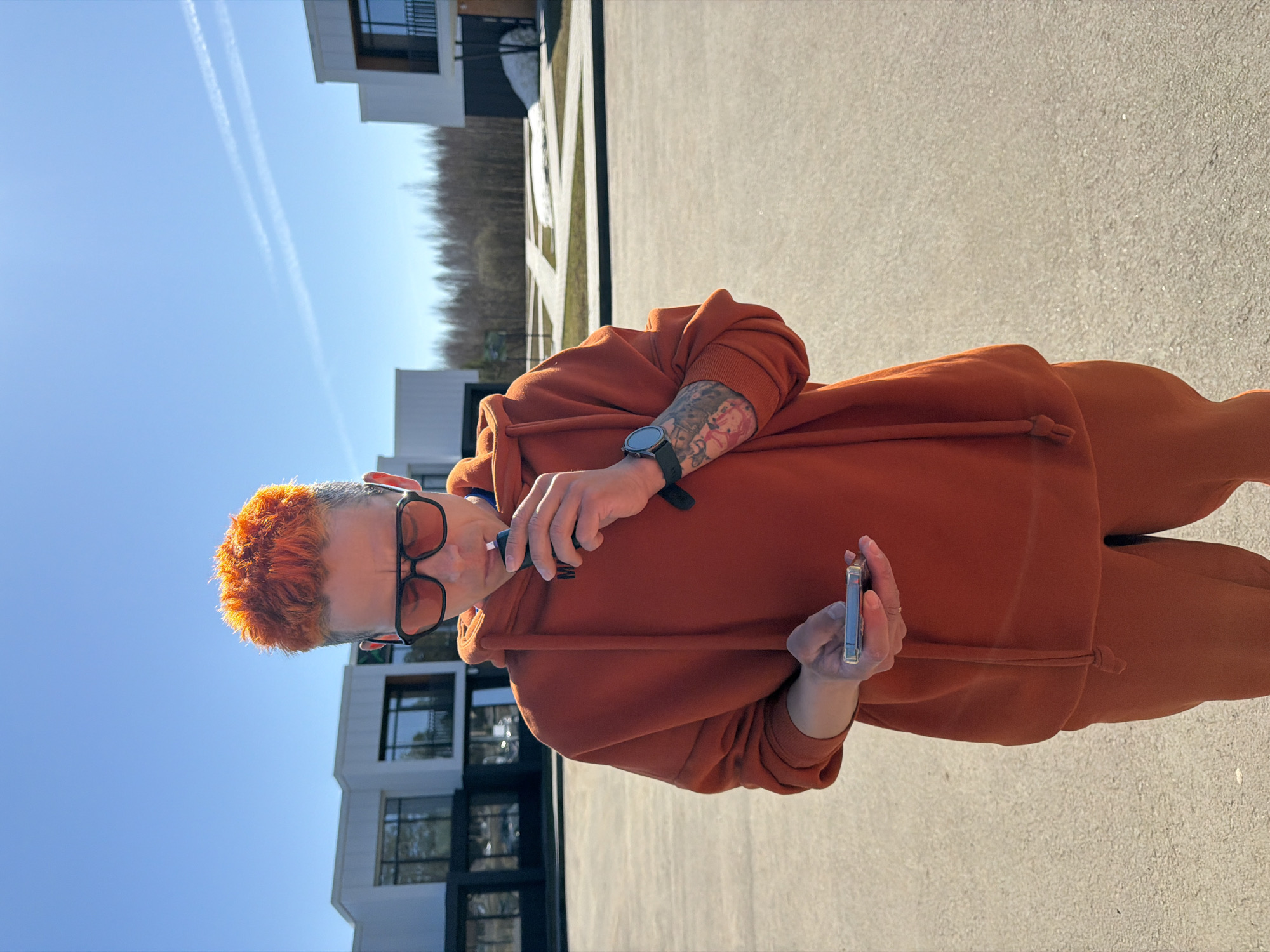 A person with orange hair and glasses, wearing an orange hoodie and pants, stands outside on a paved surface. They hold a small electronic device in one hand and a vape pen in the other. The person has a tattoo on their arm and wears a watch. In the background, there are modern buildings, a row of trees, and a blue sky with contrails. The location is Votkinsk, Russia.