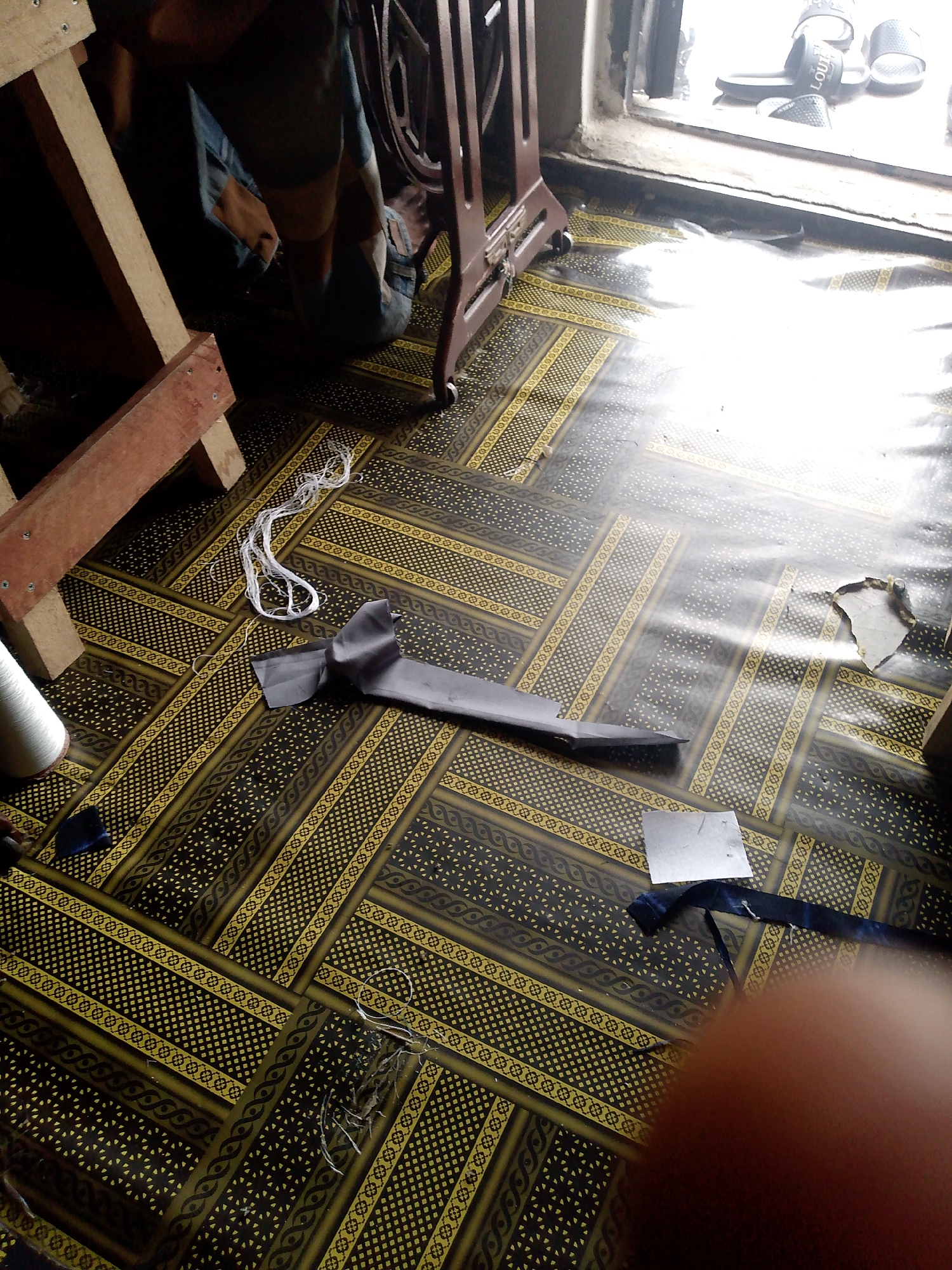 The image depicts an indoor scene with a patterned floor. A person, partially visible and wearing denim overalls, is kneeling near a sewing machine that is partially obscured. Several pieces of dark gray fabric are laid out on the floor, with one piece folded into a bow-like shape. A spool of white thread and a tangled white string are also present on the floor. To the right of the frame, a window is visible, and on the windowsill, several pairs of flip-flops are scattered. The lighting suggests it is daytime, with sunlight streaming in from the window, creating reflections on the floor. There is a wooden structure on the left side of the image. The location is identified as Jauro Yinu, Nigeria.