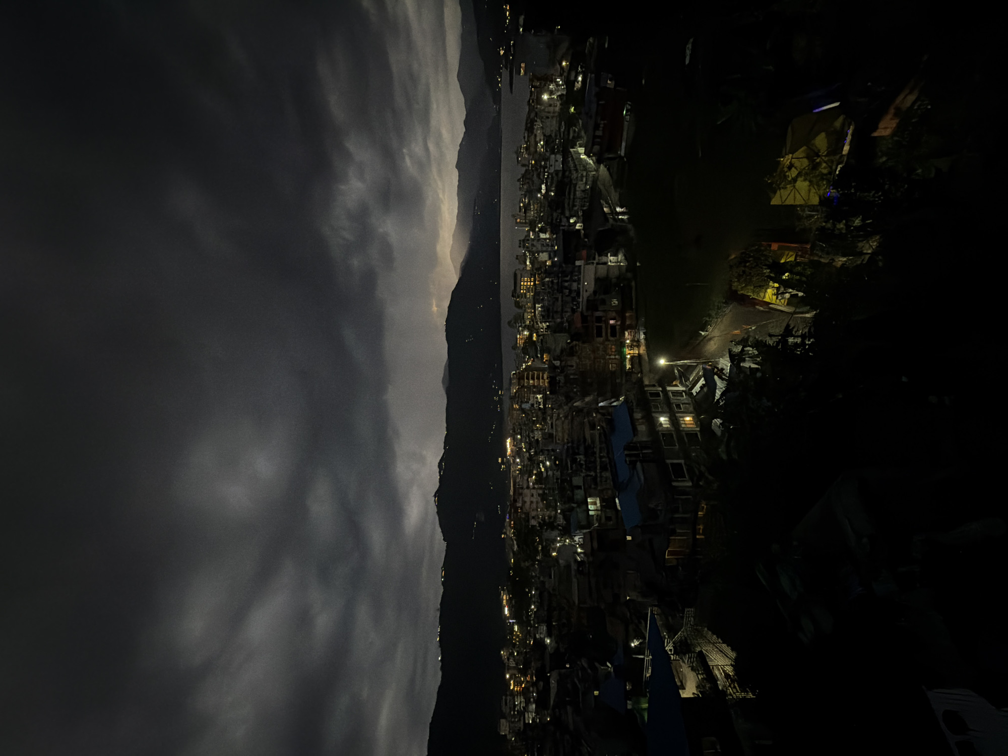 The image depicts a nighttime view of Pokhara, Nepal, likely taken from an elevated position. A mountain range divides the scene. To the left of the mountains, dark, cloudy skies dominate. To the right, the city is visible, characterized by clusters of buildings illuminated by streetlights and lights from within the structures. The darkness obscures details of the buildings. Some vegetation can be observed in the right foreground. No people are clearly visible in the image.