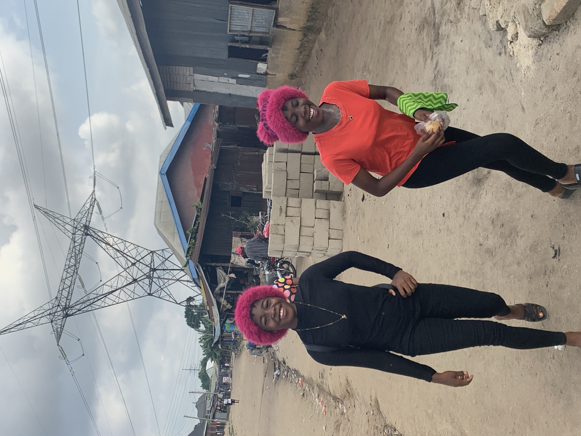 The image depicts an outdoor scene on an unpaved street in Umuode, Nigeria, under an overcast sky. In the foreground, two young females are walking towards the viewer, both smiling broadly.
The female on the left wears a black long-sleeved top, black leggings, and black slides. She has short, curly, bright pink hair (likely a wig) and a thin gold chain necklace with a small pendant. Her left hand is on her hip, and her right arm is slightly forward.
The female on the right wears an orange short-sleeved t-shirt, black leggings, and black wedge slides. She also has short, curly, bright pink hair, styled with two distinct bun-like structures on top (likely a wig). She wears a small gold chain necklace. Her hands hold a clear plastic bag containing a light-colored object, possibly food, and a folded green and white chevron-patterned cloth.
The unpaved road surface is dirt with some visible debris and loose rocks. To the right of the subjects, a stack of grey concrete blocks is present in the mid-ground. Behind the subjects, various structures line the street. These include single-story buildings with corrugated metal roofs, some with painted surfaces (e.g., a red building with a blue trim roof). A motorcycle is parked in the mid-ground near some of these structures, with a person in a red cap standing nearby. Further down the street, other individuals are visible. Dominating the upper background is a large metal electricity transmission tower/pylon with multiple power lines extending across the frame.