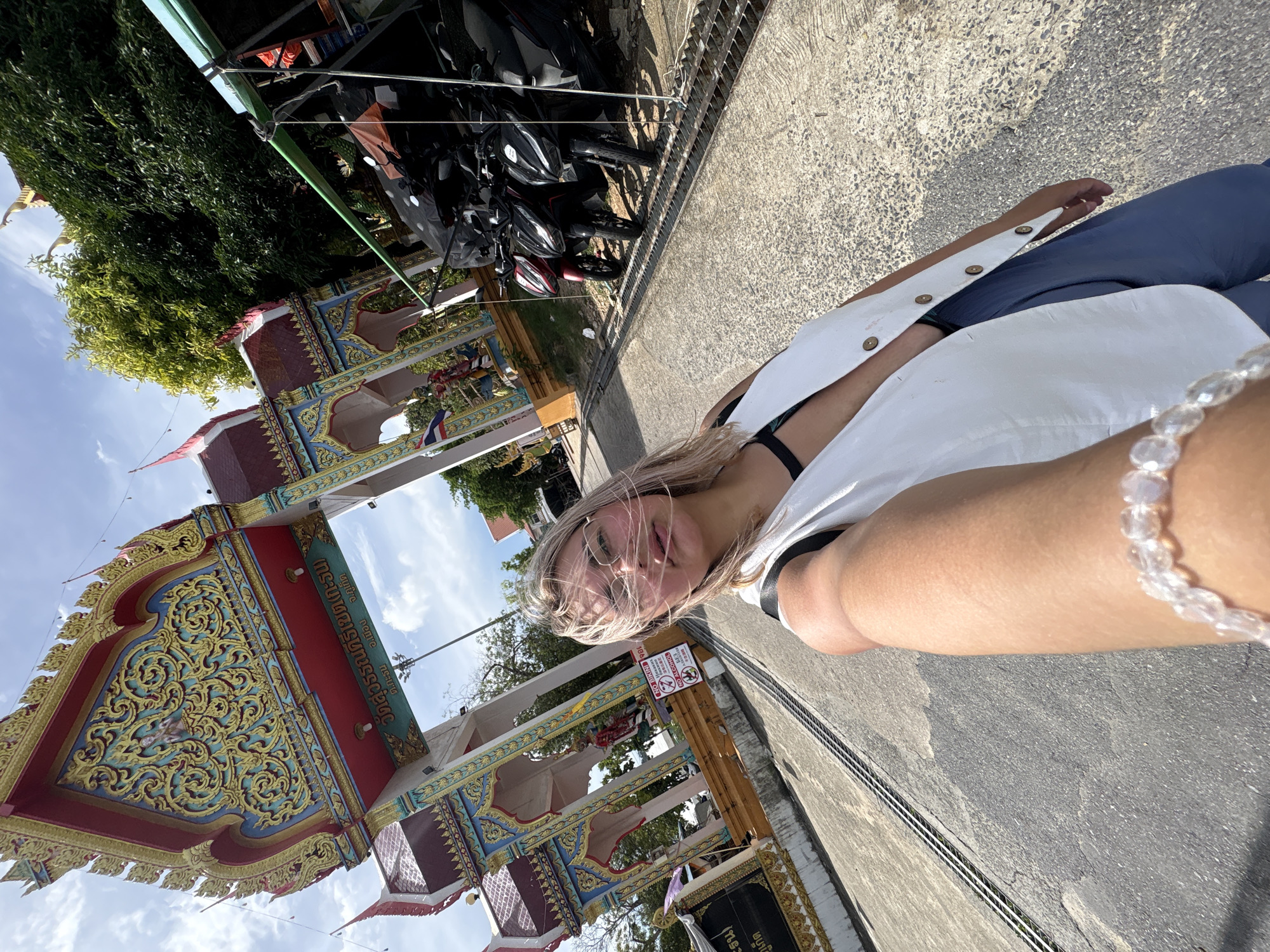 In Karon, Thailand, a woman is taking a selfie outdoors in front of a temple gateway. The gateway is ornately decorated with gold, red, blue, and green detailing, featuring Thai script and intricate carvings. A row of parked motorcycles is visible to the right of the gateway, under a green awning. The woman is wearing a white vest over a dark top and blue pants, with glasses on her face. Her hair is blonde and windswept. A beaded bracelet is on her wrist. The ground is paved with asphalt, and a drainage grate runs alongside the road. Trees and a cloudy blue sky are present in the background. Signs with text and symbols are posted near the gateway.