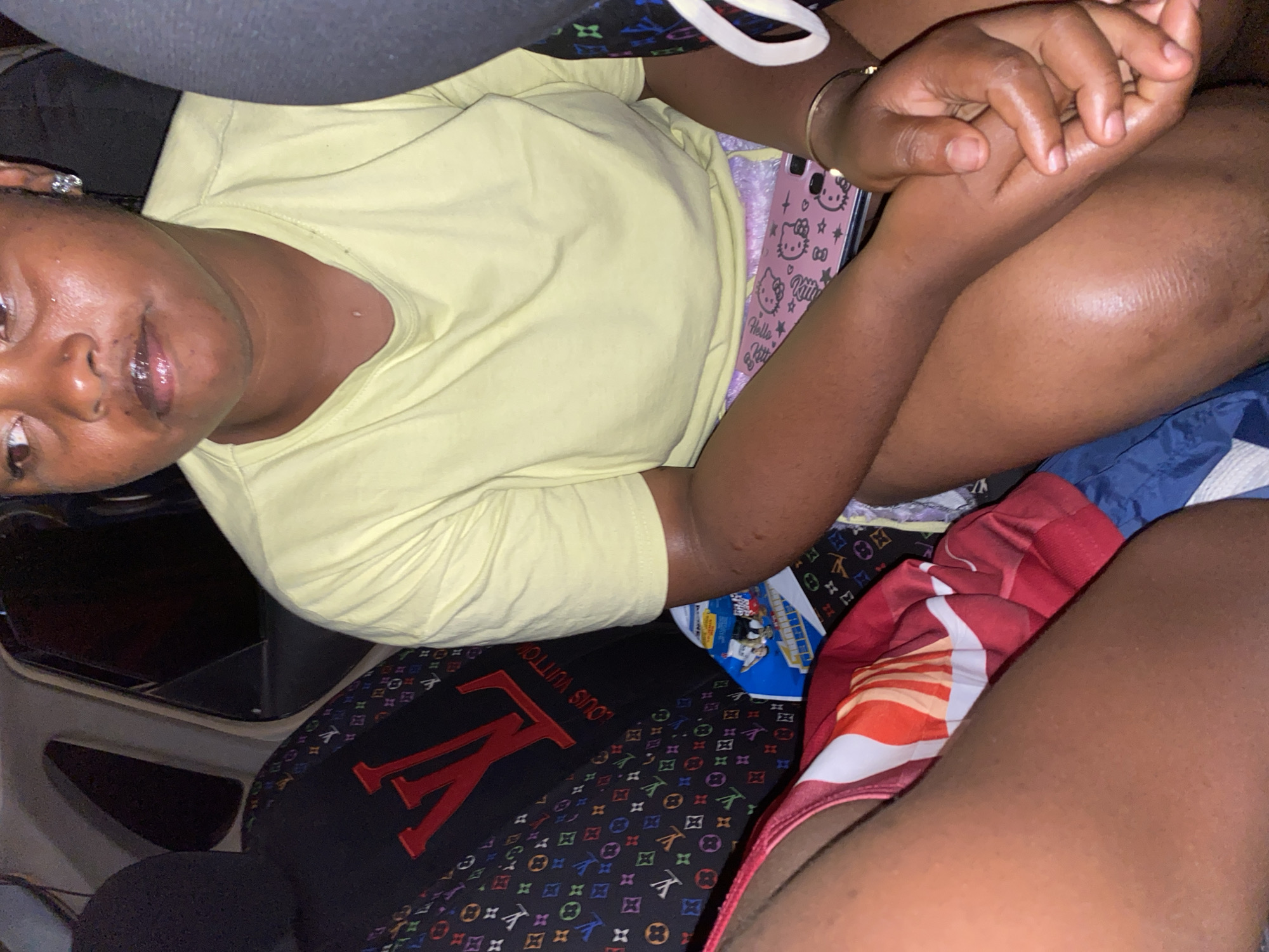 This media file shows a person, appearing to be a young Black female, sitting in a car. She is wearing a light yellow t-shirt and appears to be holding hands with someone else, though the other person is not fully visible. The background shows the interior of the car, including a black car seat with a prominent "LV" logo and a colorful pattern, possibly a seat cover or bag. There is also a pink phone case with Hello Kitty graphics visible. The person has rings on her fingers and a bracelet on her wrist. The lighting suggests it is daytime, and there are no obvious indicators of the weather. The overall mood appears relaxed and casual. The visible text includes "LOUIS VUITTON" and "Hello Kitty".