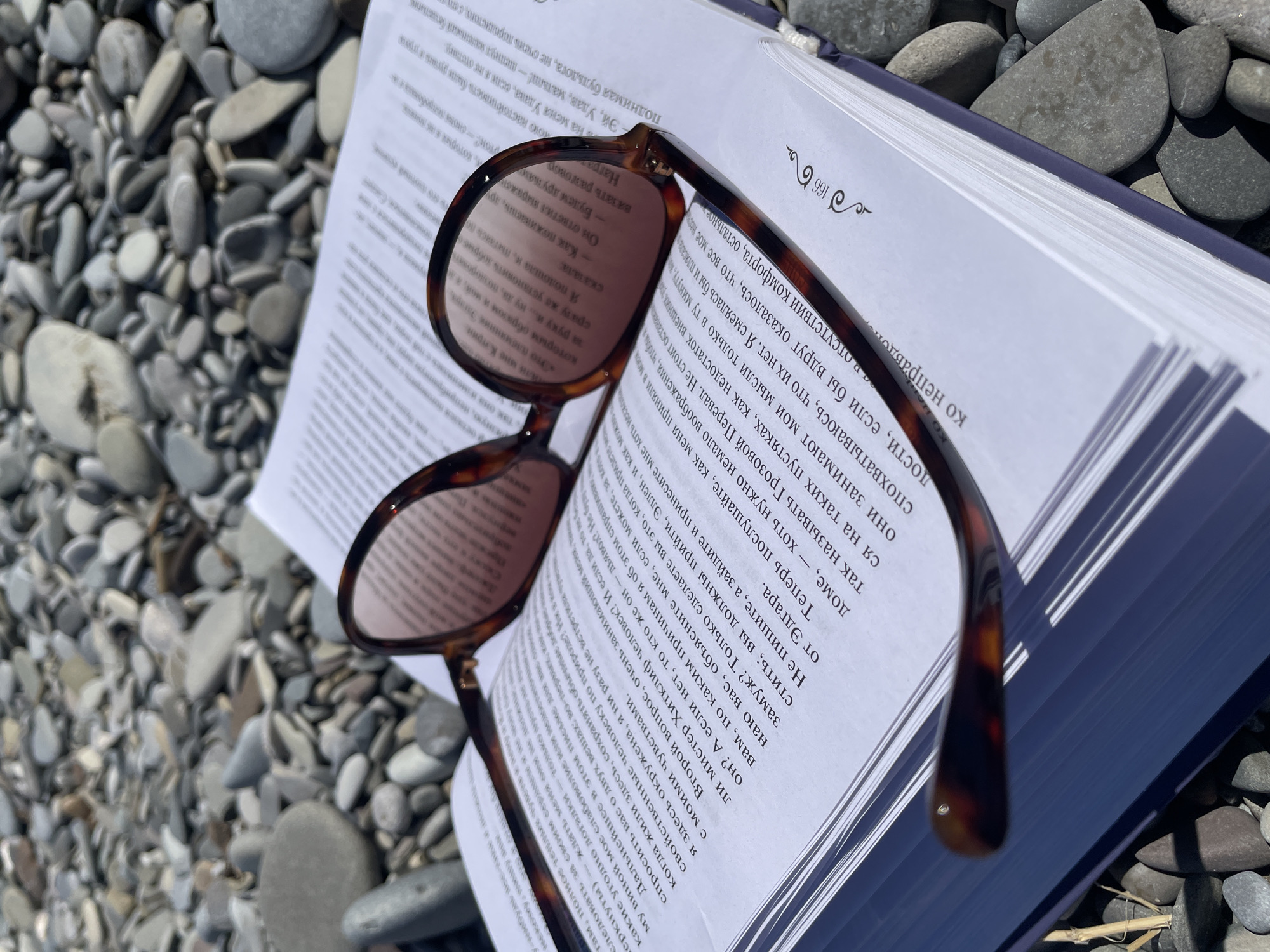 An open book with a pair of tortoise-shell sunglasses lies on a bed of smooth, grey, and white pebbles on a sunny day. The book's pages are white with black text. The sunglasses are positioned so that their lenses rest on the open pages. The background consists entirely of the pebbled surface, suggesting an outdoor setting such as a beach or waterfront. The lighting indicates bright daylight, casting subtle shadows. There are no people or animals visible in the image.