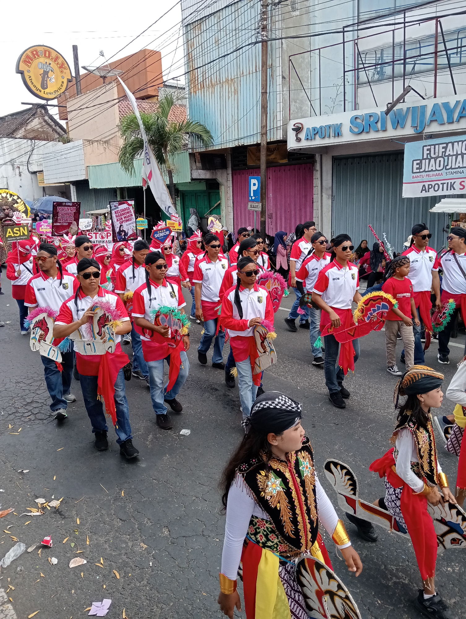 In the city of Demak, Indonesia, a parade is taking place on a paved street. A group of people, mostly young men and some children, are marching in coordinated red and white outfits. Many are wearing black headbands and sunglasses, and some are carrying colorful, horse-shaped props. The street is lined with buildings, including a business with a sign that reads "APOTIK SRIWIJAY." Some of the buildings appear to be shops or offices. There are also banners and signs visible, suggesting a community event or celebration. The ground is littered with some debris, including fallen leaves and pieces of paper. The overall atmosphere appears festive and organized.