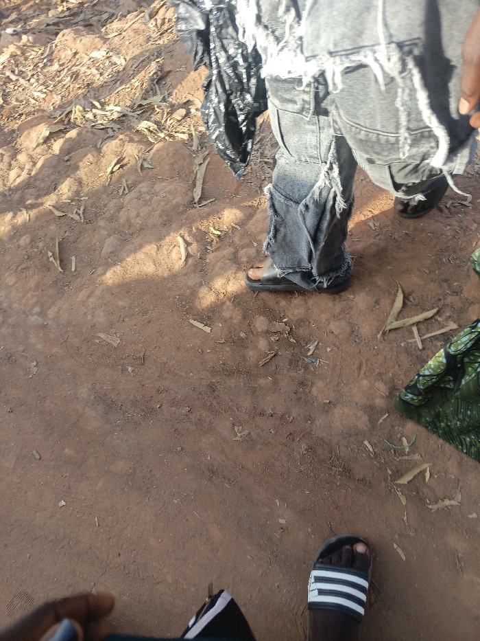 A view of ground covered in dry leaves. Someone stands in ripped jeans and sandals on the ground. Nearby, a black plastic bag and a green patterned fabric sit on the ground. In the bottom right corner is a foot in Adidas sandals. The ground is red-brown and textured. Sunlight casts shadows across the scene.