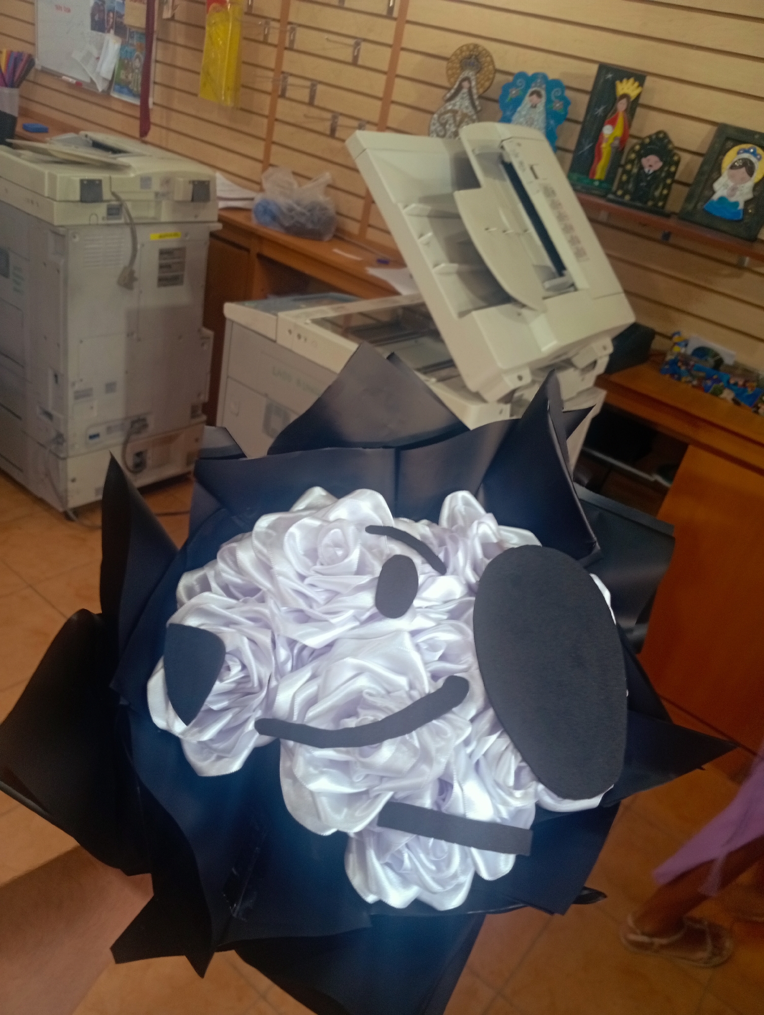 The image depicts the interior of a commercial establishment, possibly a print shop or office supply store, in Juan Griego, Venezuela. In the foreground, a bouquet of white satin roses is prominently displayed, wrapped in black paper. A black cutout in the shape of Mickey Mouse's head and ears is attached to the bouquet. Behind the bouquet, two large white office copiers are visible. The wall behind the copiers is made of wooden slats. On a shelf above the copiers, several religious icons and figurines are displayed, including depictions of the Virgin Mary. A desk with a wooden top and drawers is located to the right, with some items on it, including a blue plastic bag filled with small dark objects. The floor appears to be tiled. The lighting suggests an indoor environment, likely during daylight hours.
