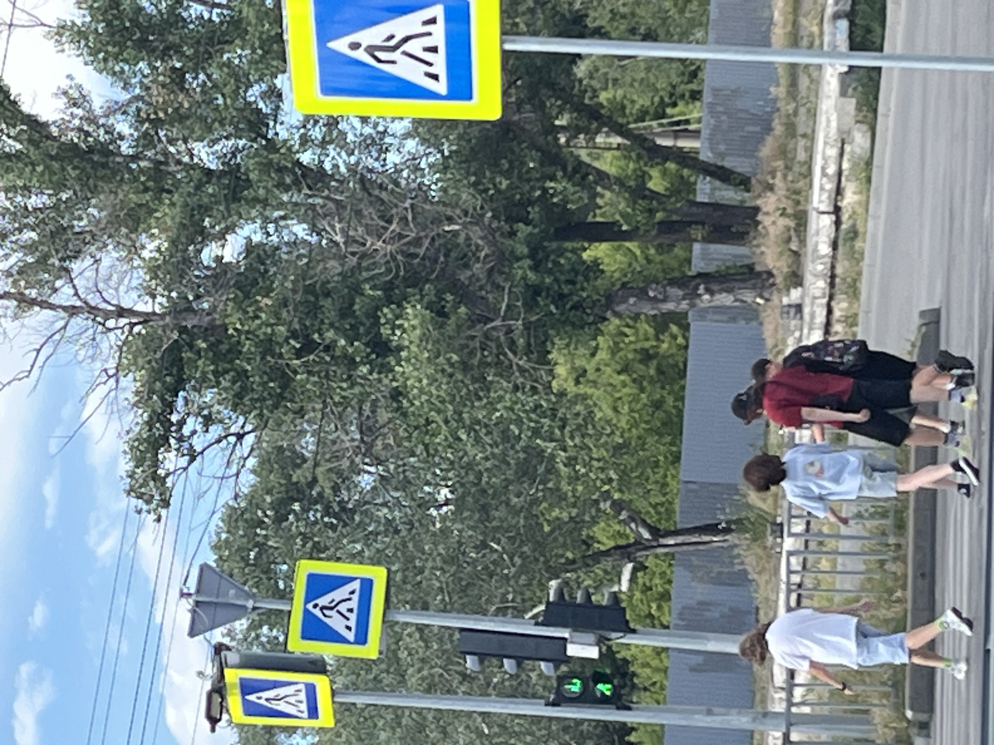 The image depicts a street scene with pedestrian crossing signs and a traffic light in Dnipro, Ukraine. Four individuals are walking across a street. The person on the far left is wearing a white t-shirt and light blue shorts. Next to them, another person is wearing a light blue t-shirt and light blue shorts. To their right, a third person is wearing a red t-shirt and black shorts, carrying a backpack. The fourth person is partially visible on the right, wearing dark clothing. The environment includes trees, a fence, and overhead power lines. The traffic light shows a green signal.
