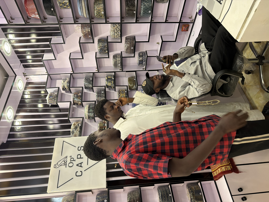 This image captures an interior scene within a cap retail establishment in Gandu Albasa, Nigeria. Three adult males are present. The shop's walls feature white, staggered display shelves showcasing numerous caps, many individually encased in clear plastic wrapping, interspersed with black and white vertical striped panels. A prominent white sign bearing the black logo "Op⁶ CAPS" and an owl illustration is affixed to the left wall. Overhead, recessed spotlights illuminate the space.
On the left, a male individual in a red and black plaid short-sleeved shirt and black track pants holds a string of light-colored prayer beads (tasbih) in his right hand. Adjacent to him, a second male, dressed in a long white traditional robe, stands looking downwards, with his right hand positioned near his chin. To the right, a third male, bearded and wearing a light grey long-sleeved shirt, black pants, and a black baseball cap, is seated in a black office chair, which is covered with clear plastic. He is actively engaged with a dark smartphone held in both hands, an earbud wire visible. A white laptop is partially visible on a counter surface to his right. A red and yellow patterned prayer mat lies partially visible on the floor in the lower left quadrant.