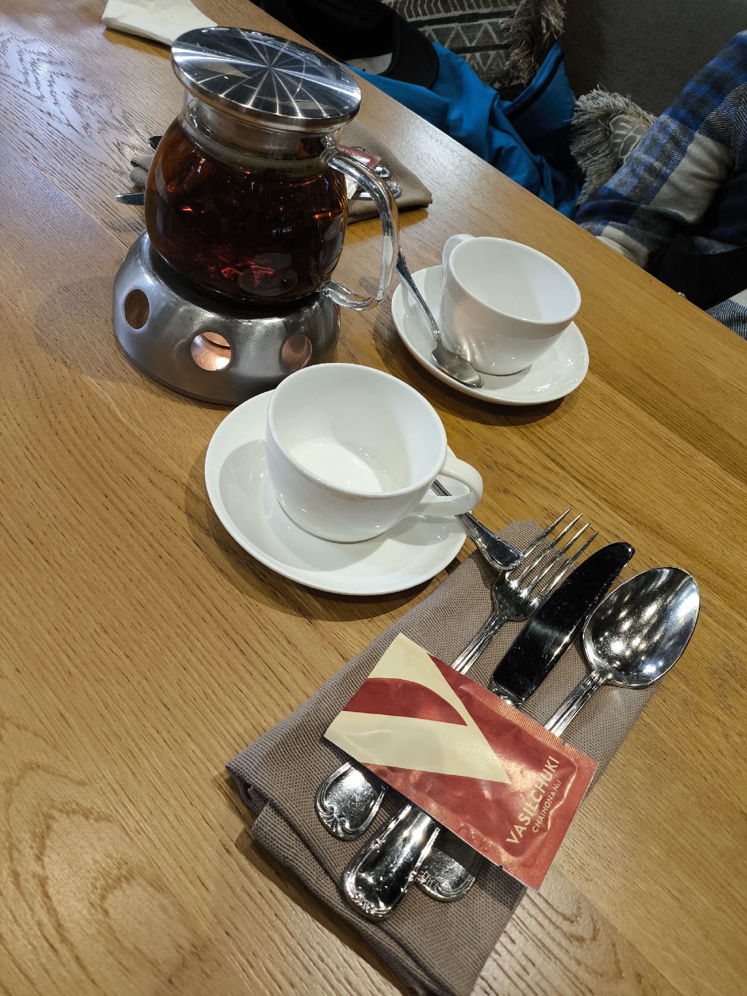On a wooden table, there is a tea service set up for two people. The teapot, filled with dark liquid, is placed on a metal warmer. Two white teacups sit on white saucers. Each cup has a small spoon. A gray napkin with a set of cutlery (spoon, fork, and knife) is positioned near one of the cups. Resting on the napkin is a small packet with the inscription "VASILCHUKI CHAIHONA No 1". This suggests that the location might be Khimki, Russia, where the "VASILCHUKI CHAIHONA No 1" restaurant is located.