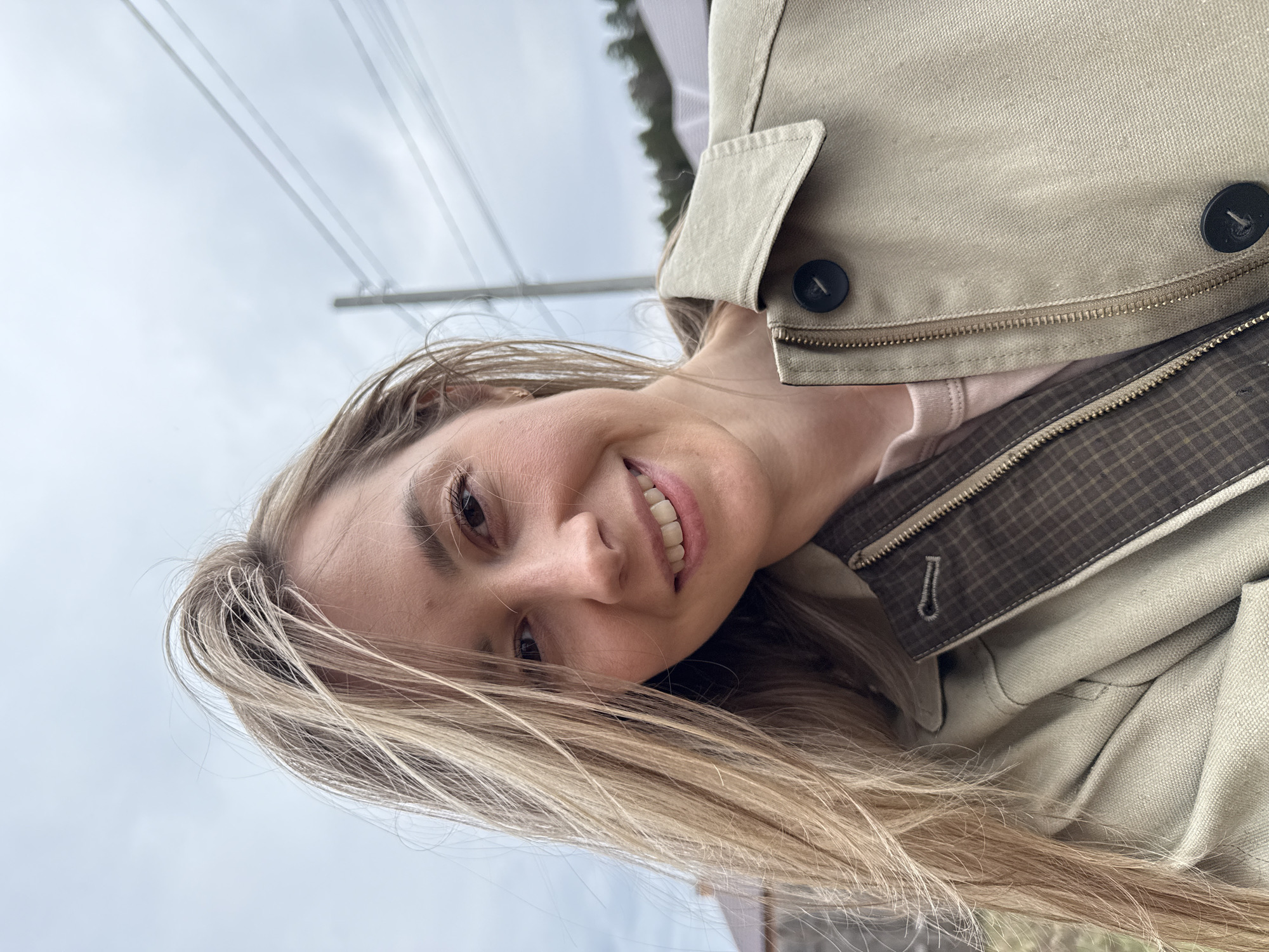 A selfie captures a smiling young woman with long, flowing blonde hair, looking directly at the camera. She has dark eyes and a bright, open-mouthed smile, revealing her teeth. She is dressed in a light beige or khaki-colored jacket, worn over what appears to be a light pink top. The jacket features dark buttons and visible zippers. One detail shot reveals a brown checkered pattern lining inside the jacket.
The background is a soft, overcast sky, indicating a cloudy or diffused light day. Thin, blurry lines, likely power lines, stretch across the sky in the upper portion of the frame. Some indistinct, dark greenery, possibly trees, can be seen blurred in the upper right corner, suggesting an outdoor location. The woman conveys a happy and relaxed emotion. There is no visible text within the image. The photo was taken in Chaikovsky, Russia.