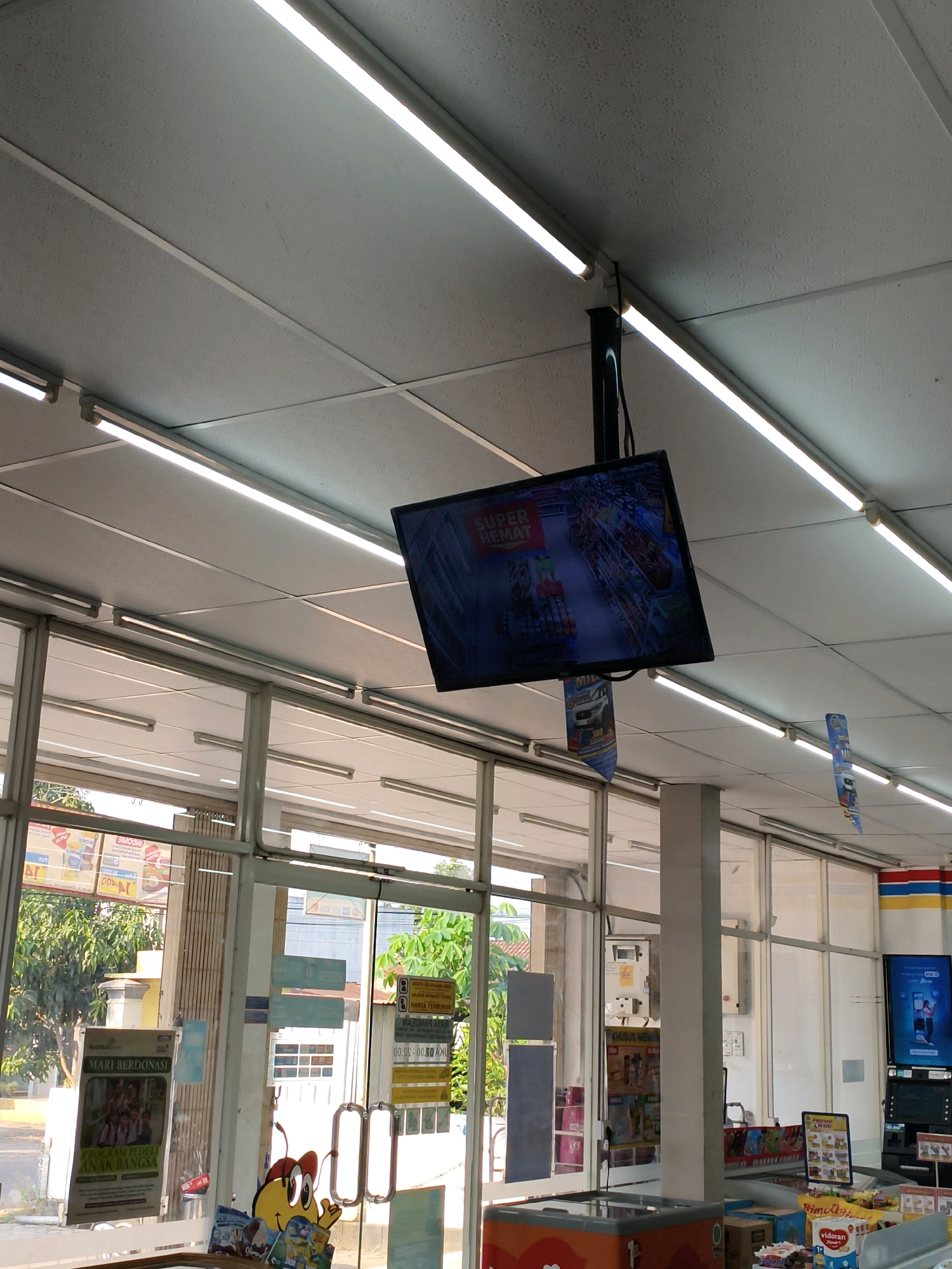 The image shows the interior of a convenience store in Sumberejo Kulon, Indonesia. A flat-screen television is mounted from the ceiling, displaying an advertisement with the text "SUPER HEMAT." The ceiling is made of acoustic tiles and is illuminated by fluorescent lights. The store has large glass windows that allow natural light to enter, revealing shelves stocked with various products. There are posters and signage on the windows and walls, including one that says "MARI BERDONASI" (Let's Donate). A refrigerator is visible in the foreground. The store appears to be clean and well-organized.
