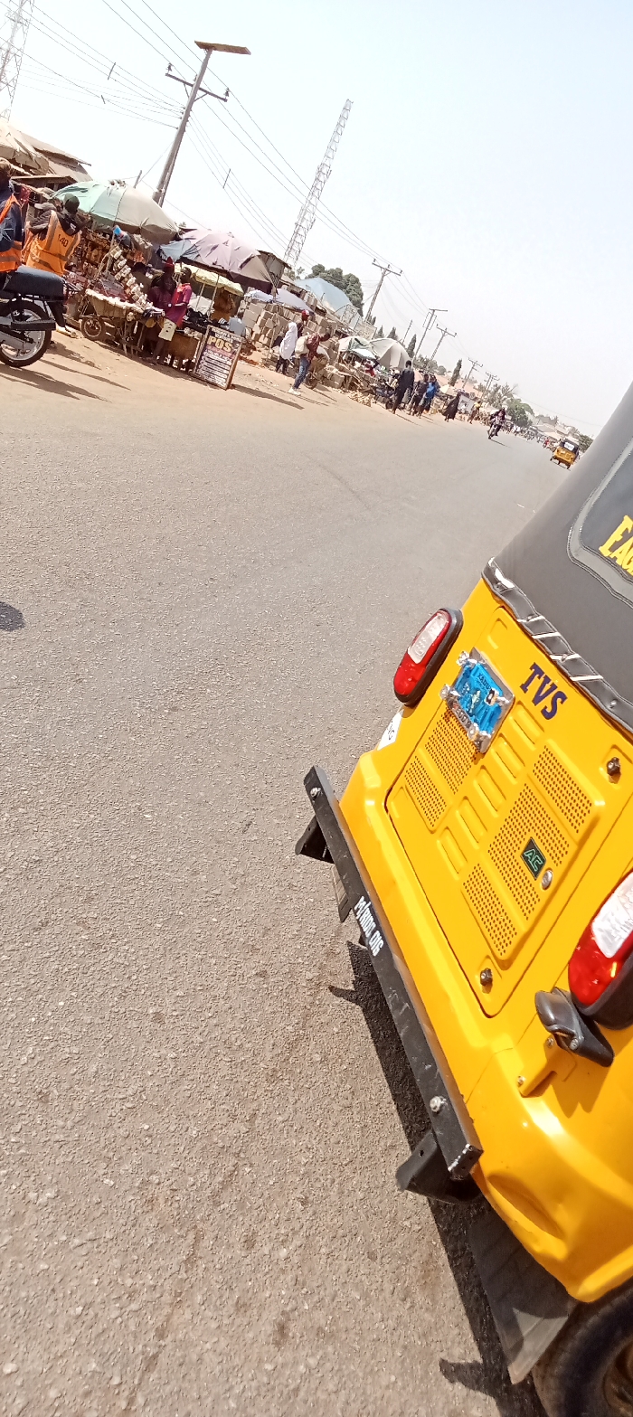 The image captures a street scene in Rido, Kaduna State, Nigeria, viewed from the rear-right perspective of a yellow TVS-branded auto-rickshaw. The auto-rickshaw features a black roof, red taillights, a black rear bumper, and a blue license plate with "KADUNA" visible. The vehicle is on an asphalt road surface.
To the left, a lively market lines the roadside, comprising multiple vendor stalls shaded by various fabric and plastic awnings, including light green, dark grey, and blue. Goods, consistent with general market merchandise and electronics, are displayed on tables and carts. A prominent "POS" (Point of Sale) service sign is visible on a standing placard. Multiple individuals are present within the market area, dressed in varied casual clothing, engaged in buying, selling, or walking. A black motorcycle is partially visible parked near the market stalls on the far left.
In the background, utility poles with power lines and a tall lattice telecommunications tower are visible against a clear, bright sky. Another yellow auto-rickshaw can be seen further down the road, receding into the distance.
