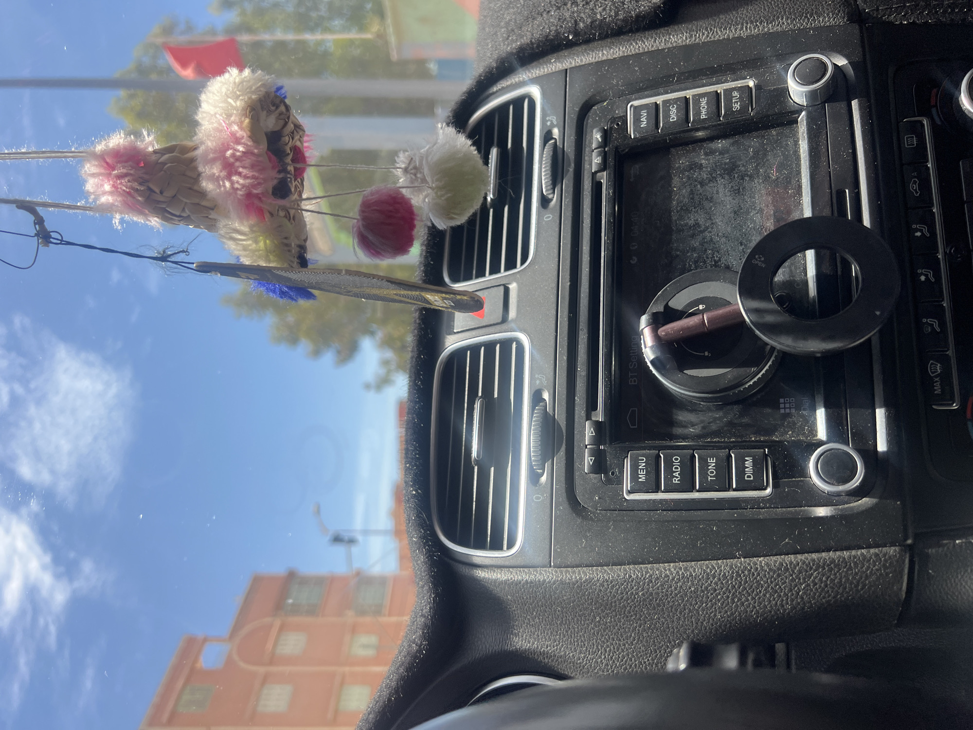 Here is a description of the image:
The image shows the interior dashboard of a car, focusing on the center console. An aftermarket car phone holder is affixed, and a car key is attached to it. Hanging from the rearview mirror is a decoration consisting of yarn and feathers. Outside of the vehicle, the sky is clear and buildings are visible, suggesting it is daytime. A partial view of an orange-colored building is visible through the windshield. Based on the building style, and the items present in the car, it is highly probable the location of this image is Ouaouizert, Morocco, since those kinds of decorations are common there.