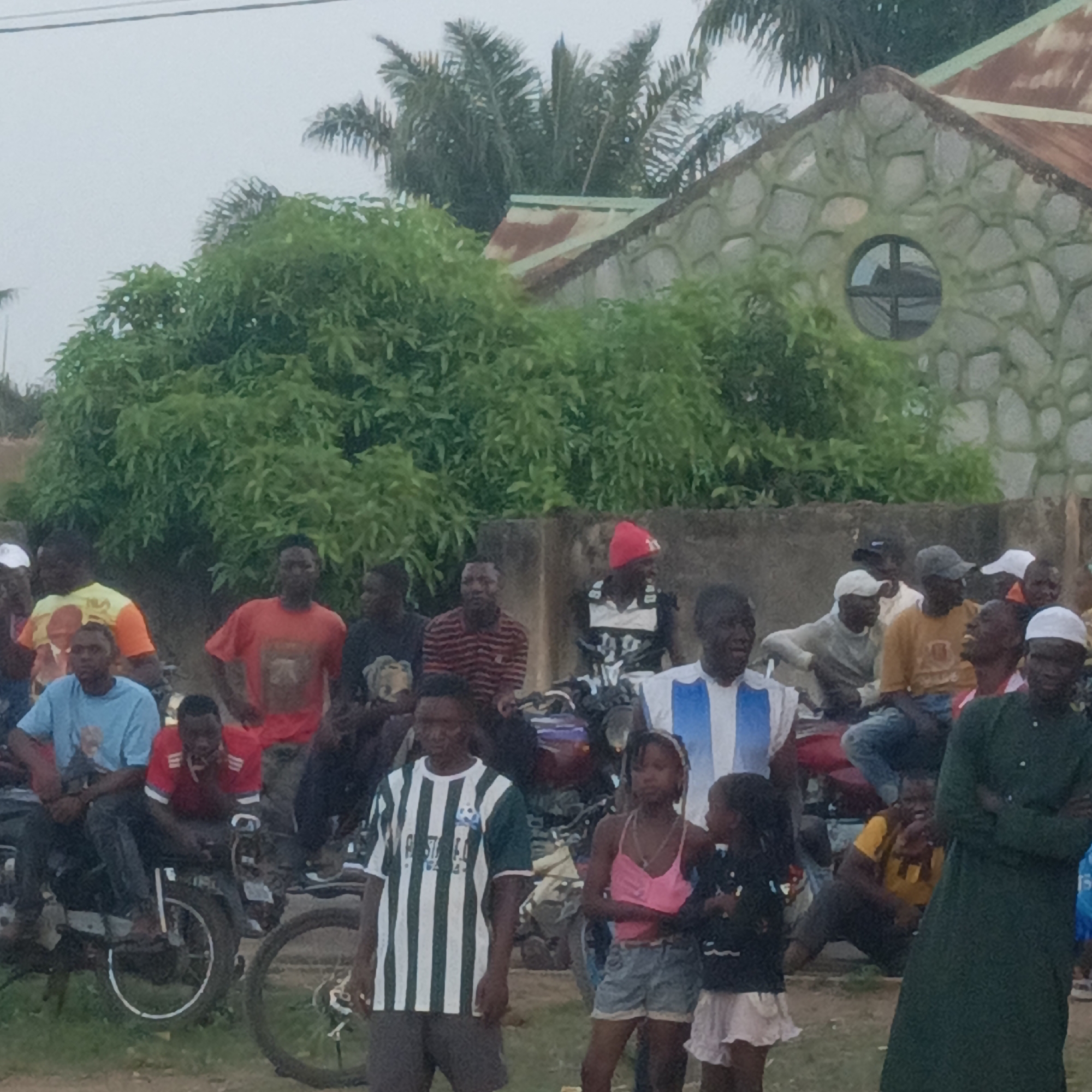 This image depicts an outdoor gathering of people in Akwanga, Nigeria. The scene is populated by a crowd, largely composed of men and a few children, standing and sitting in what appears to be a casual assembly. Many individuals are seated on or standing near motorcycles, indicating their presence or a common mode of transport. A bicycle wheel is also visible in the foreground.
In the background, there is a building with a distinctive facade made of what appears to be stone or stone-patterned material. Its roof is made of corrugated metal, showing signs of rust. A notable architectural detail on the building is a circular window with cross-shaped panes. Lush green trees, including palm trees, surround the building and form a verdant backdrop.
The time of day appears to be overcast, as indicated by the soft, diffused lighting and lack of harsh shadows. People are dressed in casual attire, including various t-shirts, some with stripes. One man is seen wearing a light blue and white striped jersey, possibly representing the Argentina national football team. Another individual wears a green and white striped jersey, likely a football club kit. A man in the foreground is dressed in a long, dark green garment, and another wears a red knit cap. The expressions of the individuals are generally neutral or observant, suggesting a relaxed social gathering or watching an unseen event. There is no clearly visible text that can be deciphered on clothing or other objects.