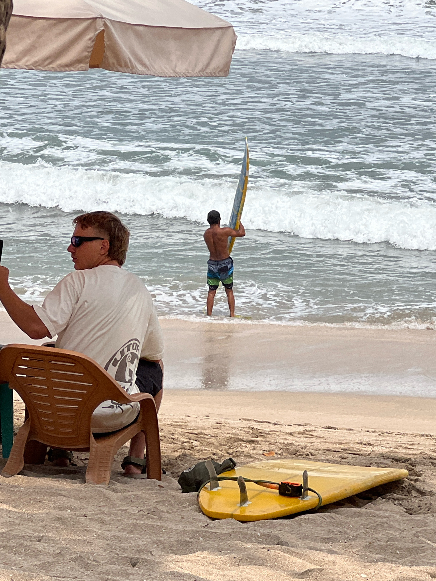 In Kuta, Indonesia, on a sandy beach, a person is seated in a brown plastic chair, facing away from the camera, holding a smartphone in their left hand. They are wearing a white t-shirt with a graphic design on the back and black shorts. The person is under a tan umbrella. In the background, a second person stands in the ocean, holding a yellow and blue surfboard vertically. This individual is wearing patterned swim trunks. A second yellow surfboard with black fins and a black leash lies on the sand in the foreground. The ocean is choppy with white waves breaking. The sky is overcast.