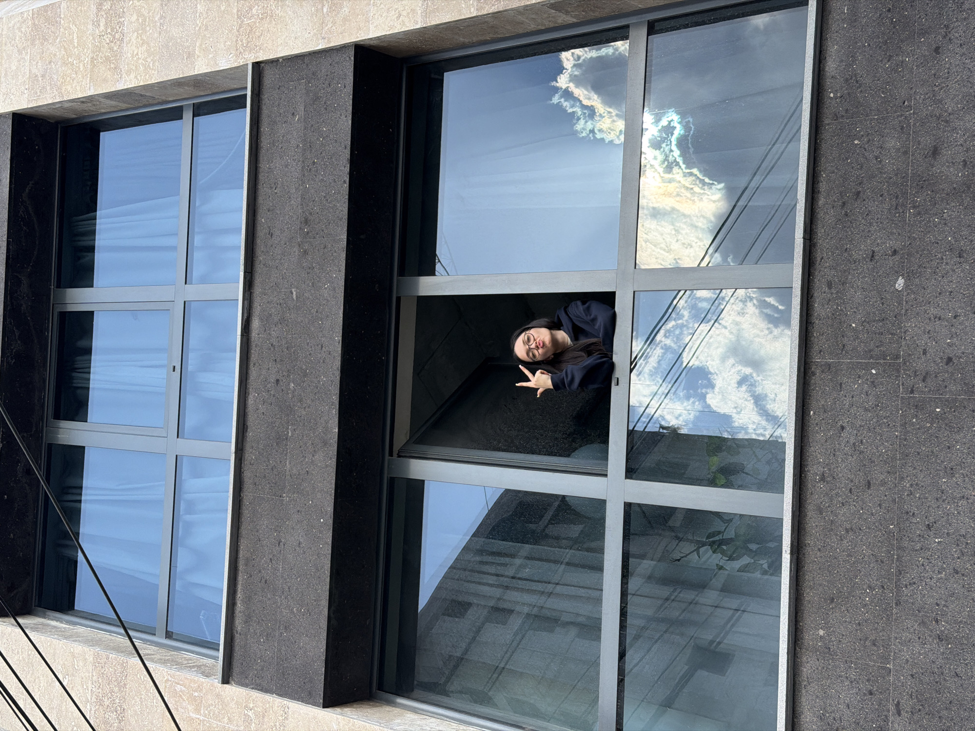 A woman is leaning out of an open window in a building in Yerevan, Armenia. The window is divided into panes and reflects the sky and clouds. The building is made of stone, with a dark stone pillar between the windows. The woman is wearing glasses and a dark top, and she is making a hand gesture. There are wires visible in the lower left corner of the image.