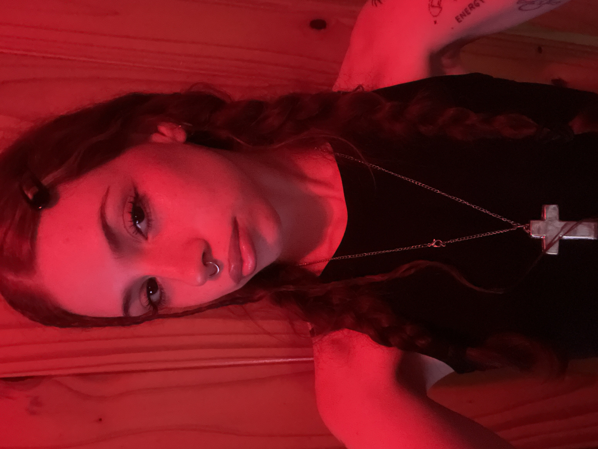 The image shows a young woman lying on a wooden surface under red lighting. She has long, wavy hair with braids, a nose ring, and a cross necklace. A tattoo is visible on her arm. The location is not discernible, so the city of Buenos Aires, Argentina cannot be verified.