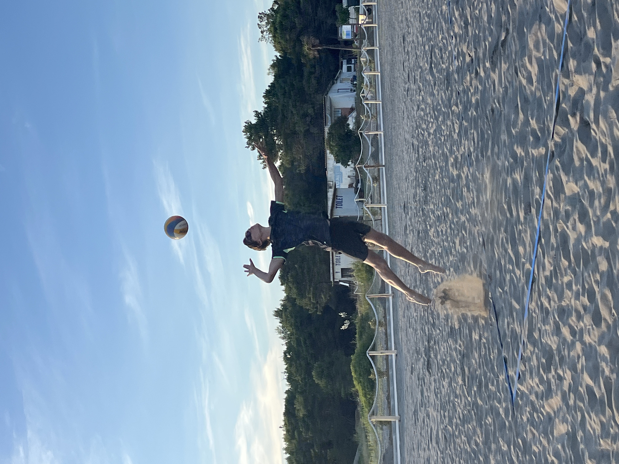 A young male volleyball player is captured mid-air during a jump on a sandy beach, serving or spiking a ball. The player is wearing a dark t-shirt with a green accent and black shorts. His body is arched backward, with one arm extended upwards towards the ball, which is visible in the upper center of the frame. His legs are bent, and his feet are off the ground, with sand being kicked up from his jump. The beach appears to be a volleyball court, indicated by a blue line visible in the sand. In the background, there is a line of trees, a small building with "TOILET" signage, and a clear blue sky with sparse white clouds. The setting suggests an outdoor recreational area, possibly a beach resort, which aligns with the known locations in Pistula, Montenegro, a region known for its coastal attractions. The lighting indicates it is likely late afternoon or early evening, with the sun casting soft light.