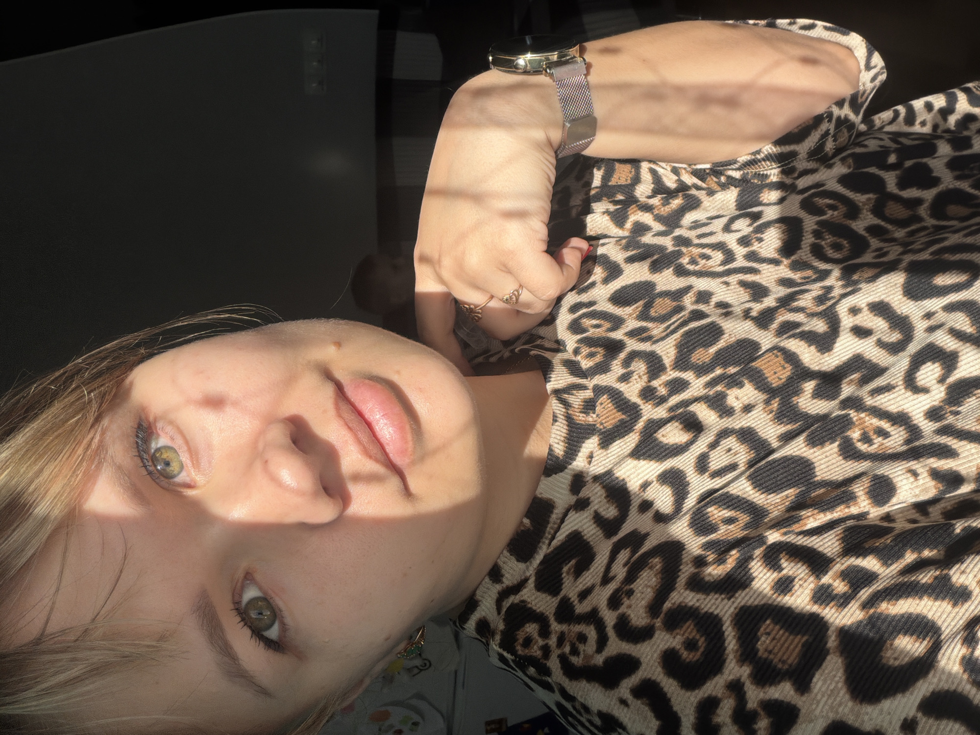 A close-up, angled selfie depicts a fair-skinned woman with light brown hair and green eyes. She is wearing a leopard-print top and a silver and gold wristwatch. The woman's face is partially illuminated by sunlight, creating shadows across her face. She has rings on her fingers. The background includes a dark surface, possibly a desk or table. The location is Chaikovsky, Russia.