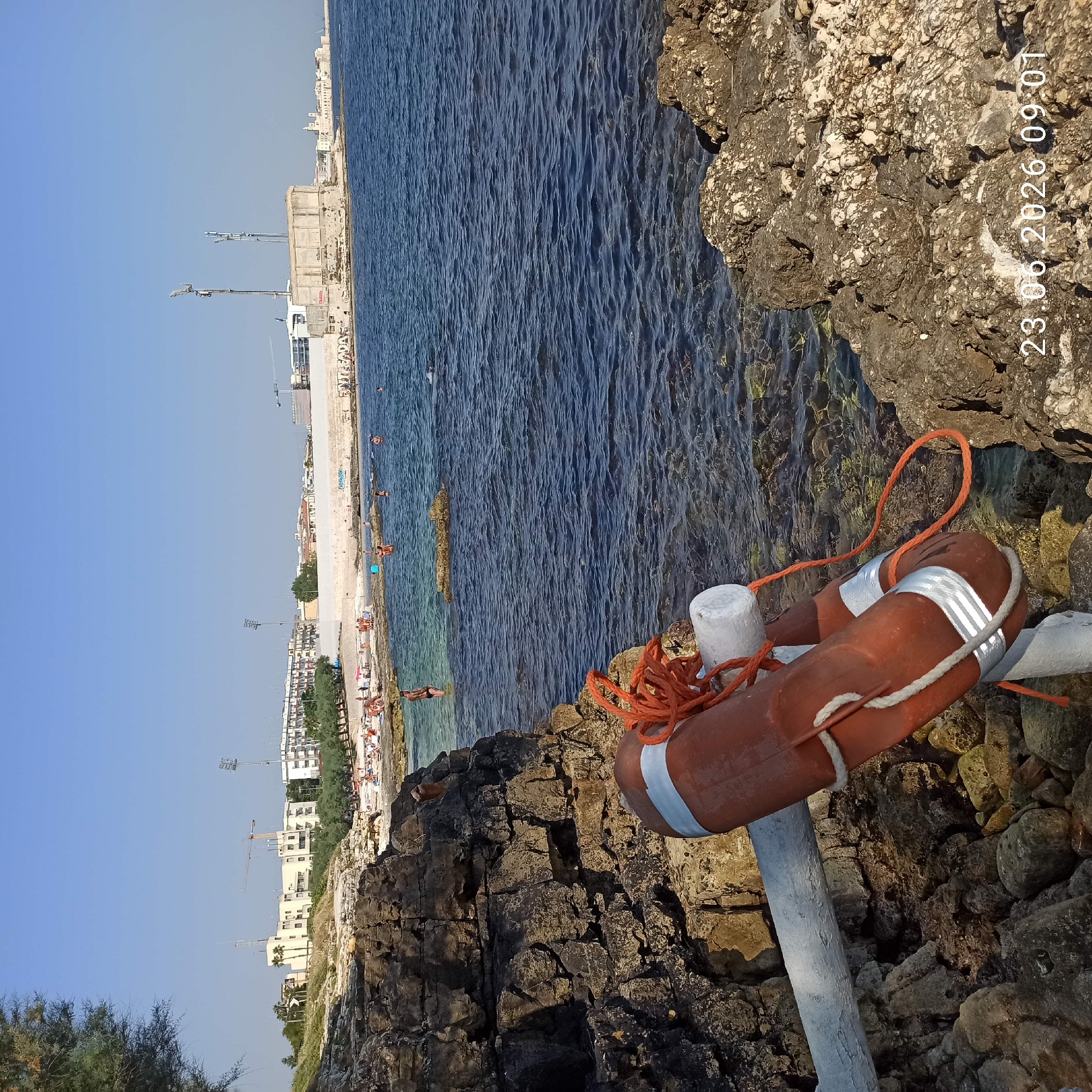 The image depicts a scenic coastal view with a clear blue sky and calm sea. The foreground is dominated by rocky terrain and a life preserver with orange rope, attached to a white pole. In the background, a beach lined with buildings and trees stretches along the coastline, with a few people visible on the beach and in the water, suggesting a leisurely day. The presence of construction cranes in the distance indicates ongoing development in the area. The lighting suggests it is daytime, possibly late afternoon given the warm tones on the rocks. Visible text on the image reads "23.06.2026 09:01". The overall atmosphere is tranquil and typical of a summer day by the sea in a populated coastal town.