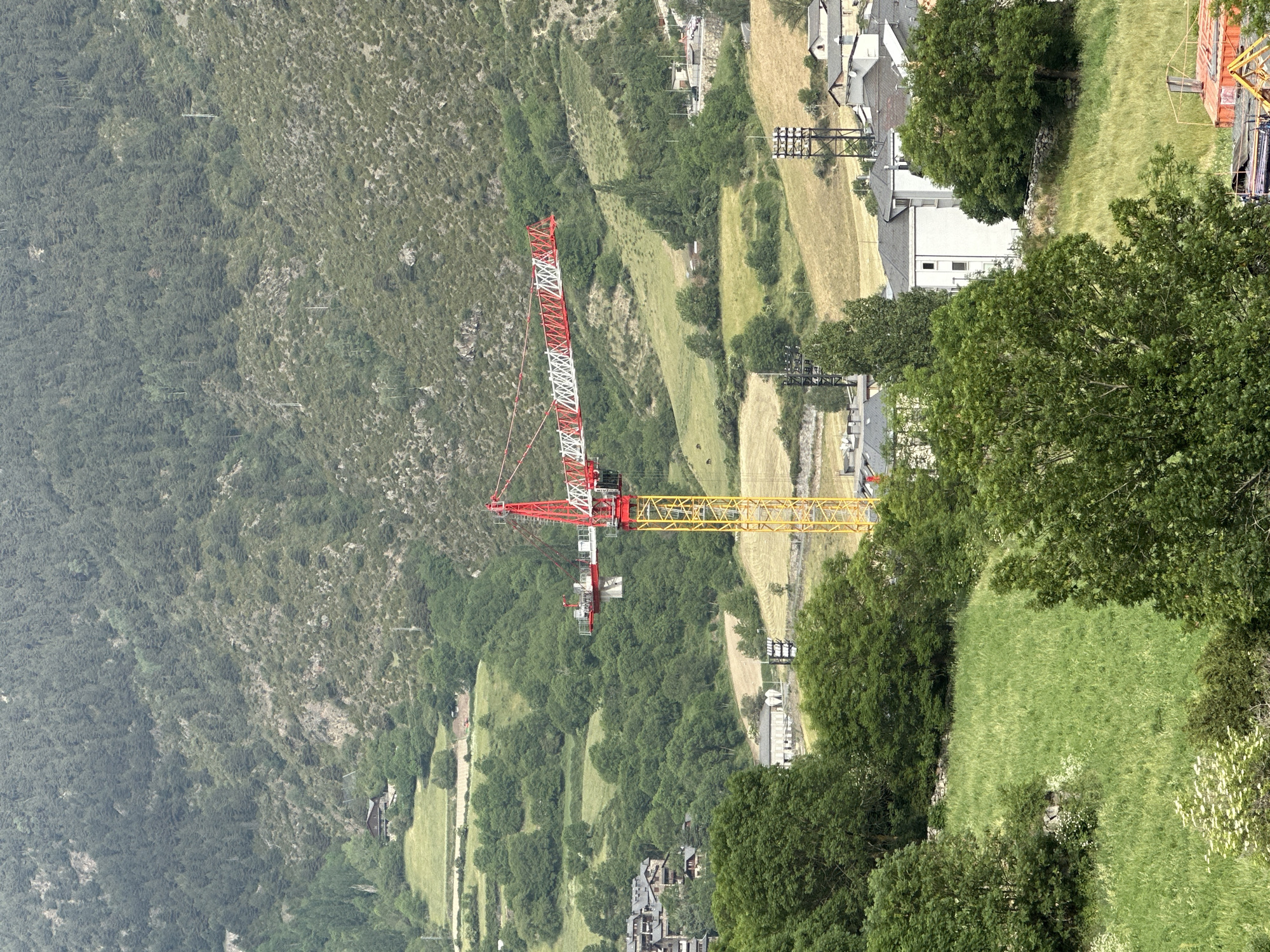 The image displays a wide, panoramic view of a mountainous valley, consistent with the landscape of Encamp, Andorra. In the mid-ground, a prominent red and white tower crane with a long yellow horizontal jib signifies ongoing construction. The crane stands against a backdrop of varied terrain.
The valley floor and lower slopes feature a patchwork of green, verdant fields and drier, yellowish-brown cultivated areas, interspersed with numerous deciduous and coniferous trees. Several buildings, primarily houses with light-colored facades and dark roofs, are visible, particularly in clusters on the right side of the valley. Utility poles and power lines are also present, indicating infrastructure.
The background is dominated by steep, densely forested mountain slopes, exhibiting deep green foliage and some rockier outcrops. The overall lighting suggests daytime, possibly under an overcast sky or with some atmospheric haze, lending a soft, diffused quality to the scene. No individuals or discernible text are visible. The image captures a blend of natural mountainous beauty and human development.