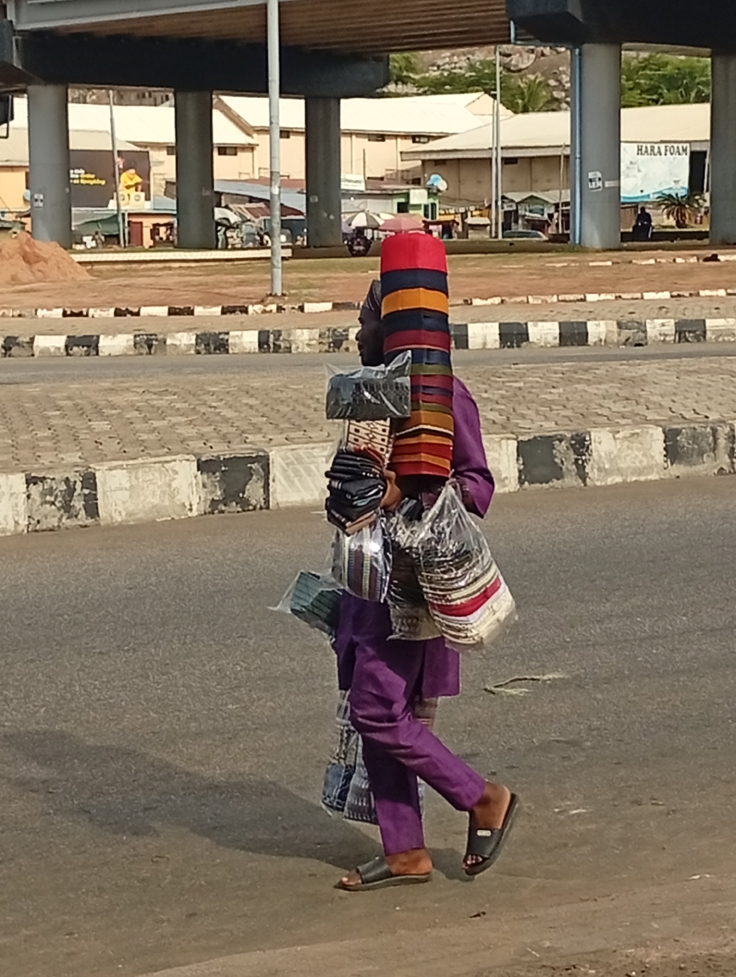 A street vendor walks along a gray road in Lokoja, Nigeria. He is wearing a purple outfit and black sandals, and he is carrying a stack of goods, including colorful hats and packaged items, both on his head and in his arms. The environment includes a sidewalk with black and white accents, buildings in the background, and a bridge structure overhead. A sign for "HARA FOAM" is visible in the background.