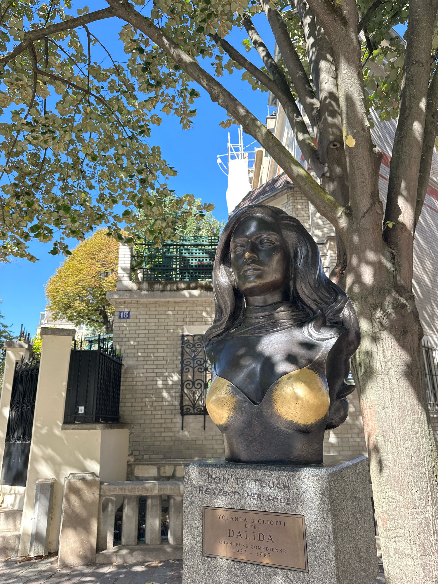 The image shows a bust of a woman on a stone pedestal, with a bronze sculpture of her upper body and head. She has long, wavy hair, and is wearing a gold-colored top resembling a brassiere. The pedestal is inscribed with "DON'T TOUCH RESPECT HER SOUL" and a plaque below reads "YOLANDA GIGLIOTTI DITE DALIDA CHANTEUSE COMEDIENNE 1933 - 1987".
The bust is located outdoors, in what appears to be a residential area of Montmartre, Paris, France, indicated by the architecture and the street number "15" visible on a brick building in the background. A large tree with dappled sunlight on its trunk and branches shades the sculpture, and a portion of a building with a white brick facade and green iron fencing is visible. The sky is clear and blue. There are no people present in the image.