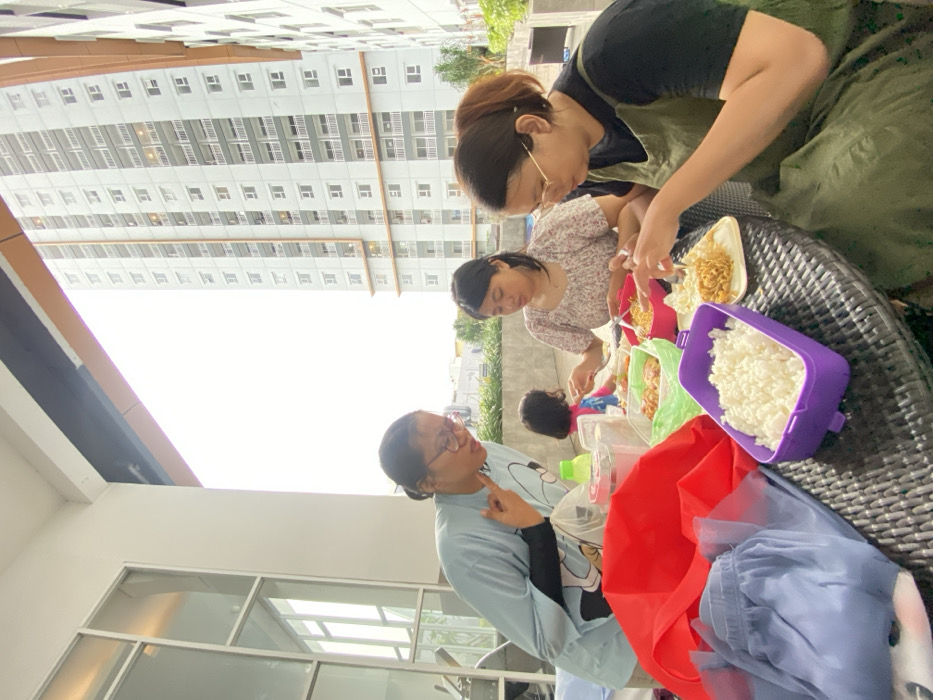 The image depicts an outdoor meal taking place on a paved terrace or balcony, identified as being in the city of Sidoarjo, Indonesia. Four individuals are visible, three adults and one child, gathered around a dark-colored, woven-texture outdoor table laden with food containers.
On the left, a woman with dark hair in glasses, wearing a light blue long-sleeved shirt, stands and leans forward, gesturing with her right hand (index and pinky fingers extended) and an open mouth, as if speaking or reacting. To her right, a second woman with dark hair, wearing a patterned short-sleeved top, is seated, looking down at a bowl of food and eating with a fork. Further to the right, a third woman with light brown hair in glasses, wearing a dark top and an olive-green apron-style dress, is seated, actively eating from a white plate with a fork. Partially visible behind the second woman is a young child in a pink top and blue bottoms, seated at the table. A hand from an unseen person is visible on the far right edge of the frame.
On the table, there is a purple rectangular container filled with white rice, a green container with what appears to be fried food, a pink bowl containing noodles, and a white plate with crispy noodles. Other items include a clear plastic container with a red lid, a green bottle, a red fabric item, and a light blue pleated skirt.
The immediate background behind the seated individuals features a low, light gray concrete wall, with some greenery beyond it. The wider background is dominated by a tall, modern multi-story apartment building, light in color with numerous balconies and windows, suggesting a residential urban environment. The sky overhead is bright white, indicating an overcast condition or overexposure. To the left, a structure with large windows and white frames is partially visible.