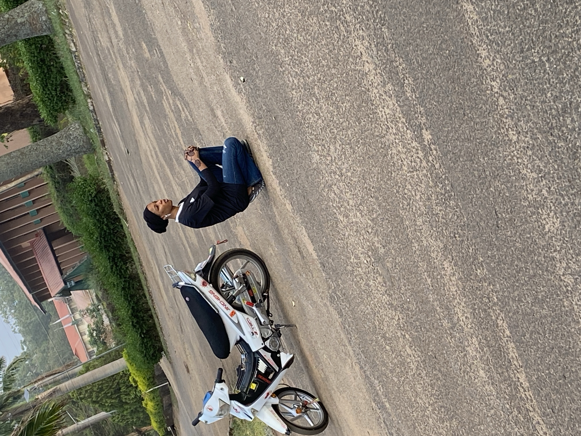 A person is sitting on a road next to a white motorcycle. The person is wearing a black shirt and blue jeans. The motorcycle is parked on the side of the road. The location appears to be Vom, Nigeria, based on the license plate number. Buildings and vegetation can be seen in the background. The road is asphalt.