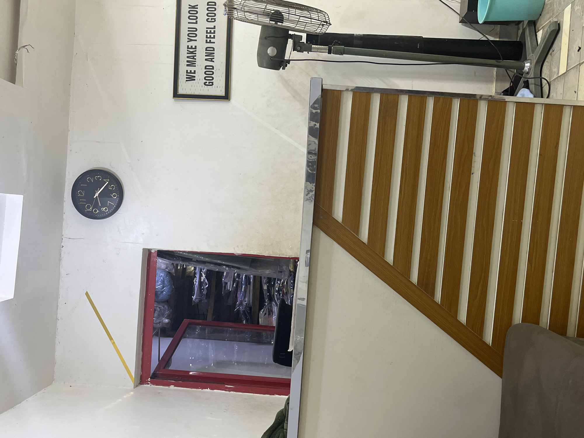 The image shows an indoor scene. On the left wall, there is a clock set to approximately 11:58, a sign that reads "WE MAKE YOU LOOK GOOD AND FEEL GOOD", and a doorway leading to what appears to be a storage or dressing area filled with clothing items hanging on racks. A yellow measuring tape is attached to the wall. To the right is a wooden staircase railing with vertical slats and a gray-colored object. A floor fan is visible towards the top of the image. The location is potentially Kubwa, Nigeria. The image contains no people.
