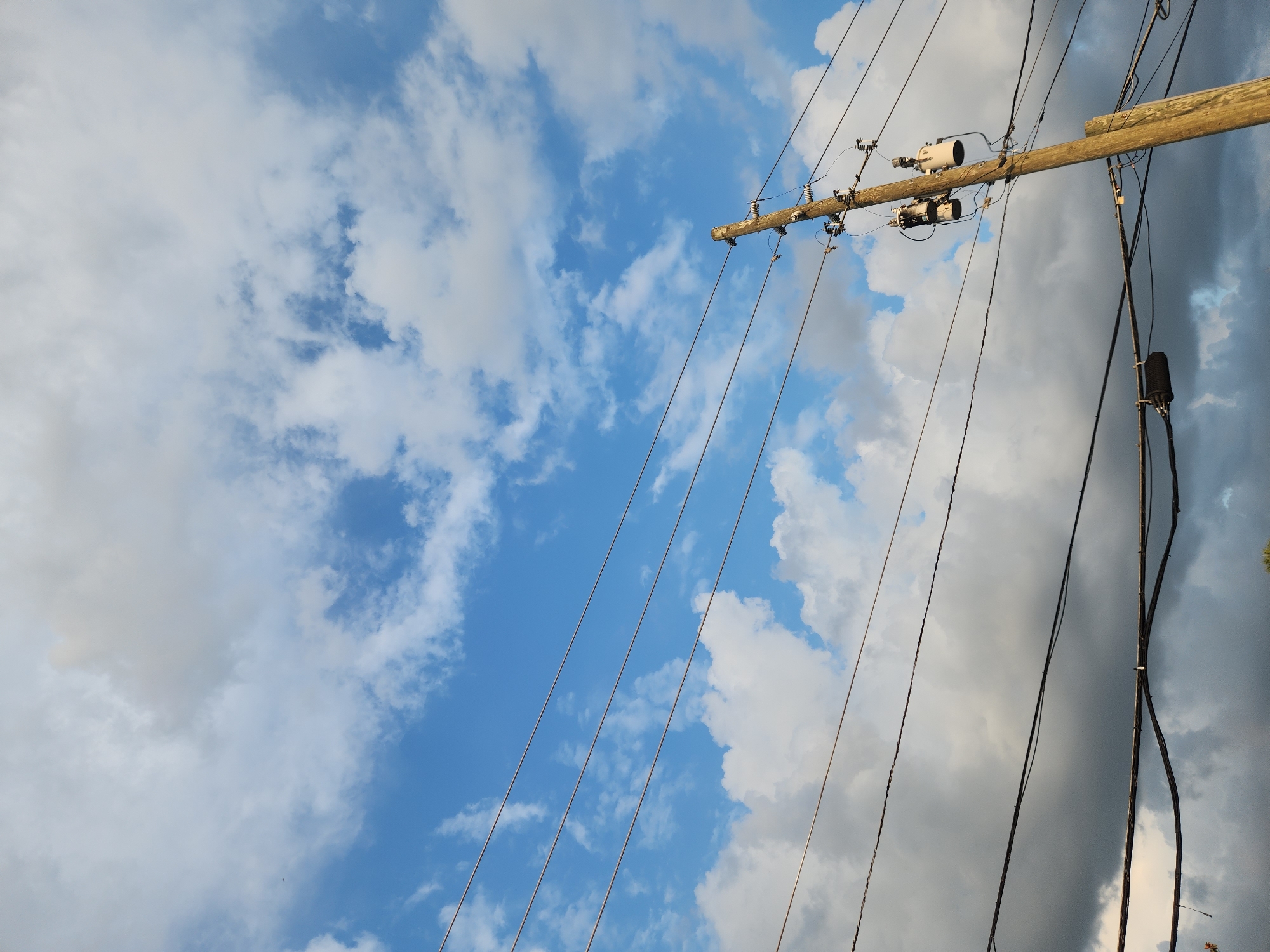 In Maitland, Florida, United States of America, a wooden utility pole stands diagonally against a dynamic sky filled with white and gray clouds. The pole is equipped with several electrical transformers and insulators. A network of dark electrical wires stretches across the frame, intersecting the pole and leading towards the viewer. The lower portion of the image shows a darker, more ominous cloud formation, contrasting with the brighter, sunlit clouds above and the clear blue sky peeking through. The scene is captured from a low angle, emphasizing the scale of the utility pole and the vastness of the sky.