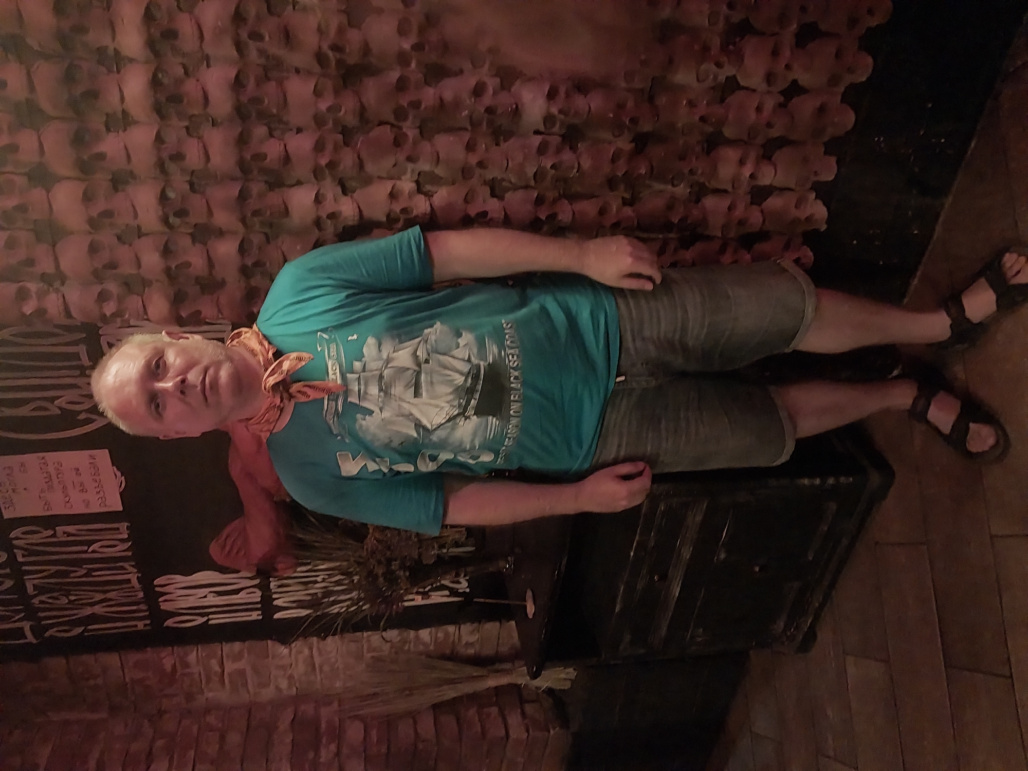 A bald, middle-aged man wearing a teal t-shirt with a ship graphic, denim shorts, and black sandals stands in front of a wall covered in numerous skull decorations. He has a light-colored bandana tied around his neck. To his left, a dark wooden cabinet with decorative metalwork is visible, and behind it, a brick wall and some dried plant material. The floor is made of wooden planks. The overall impression is of an indoor setting, possibly a bar or a themed establishment. The location is identified as Orenburg, Russia.