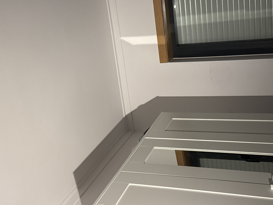 The image depicts an interior corner of a room, likely in Bragança, Portugal. The upper portion of the scene features a white ceiling with decorative crown molding along the junction with the walls. The walls are light-colored, possibly white or light grey. In the lower left quadrant, the upper section of a light-colored cabinet or wardrobe with recessed panel doors is visible. The cabinet's surface reflects portions of the room, including a dark-framed window. Above the cabinet, a prominent, elongated shadow is cast onto the ceiling and upper wall, extending from the cabinet's edge diagonally towards the upper right.
In the lower right quadrant, a window with a dark frame and horizontal blinds is present. A wooden lintel or trim piece is positioned directly above the window frame. A vertical band of bright light, potentially from another light source or a reflection, is visible on the wall to the left of the window and below the crown molding. No individuals, discernible actions, or direct interactions are visible in the static indoor environment.