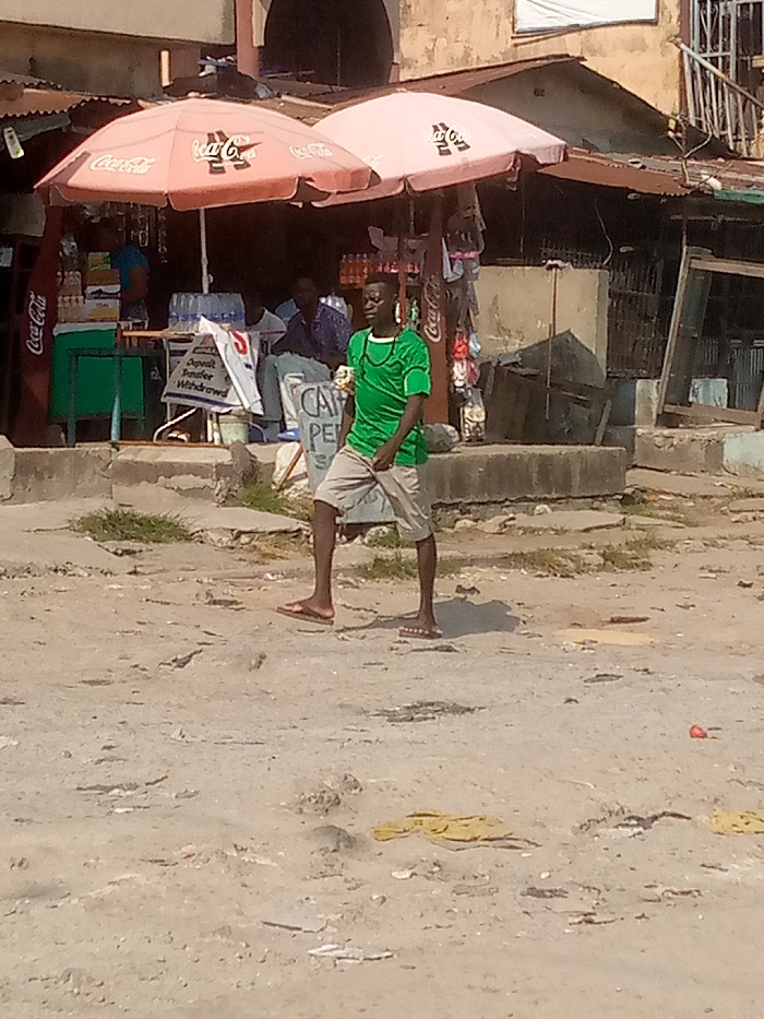 This image depicts an outdoor street scene in Badore, Nigeria, under daylight conditions.
In the foreground, an unpaved, dusty ground with uneven terrain and scattered debris, including a small red object and yellow fabric pieces, extends across the frame. A young adult male, dark complexion, is positioned slightly right of center, mid-stride, facing partially left. He wears a short-sleeved green shirt with a subtle pattern, light-colored (possibly khaki or grey) shorts, and brown flip-flops. A dark chain or strap is visible around his neck, and he holds a small, light-colored object in his right hand.
In the midground, a raised concrete curb separates the unpaved area from a retail stall or shop operating beneath two large, light-pink Coca-Cola branded market umbrellas. To the left, a dark-skinned individual in a blue shirt is partially visible behind a green counter, with various bottles and containers displayed. A Coca-Cola cooler stands upright on the far left. To the right, two dark-skinned individuals are seated; one wears a blue patterned shirt and faces left. A white sign reading "Deposit, Transfer, Withdrawal" is prominent, indicative of a financial service point. Another sign, partially visible, reads "CAMPENS" or similar. Various merchandise, including small packaged items, hangs from a display near the right umbrella.
The background consists of older, light-colored buildings with corrugated metal roofs. A dark arched doorway is visible in the central background building. A metal railing or staircase is present on the right side of a background structure. The overall environment suggests an urban or semi-urban setting with established, somewhat weathered infrastructure.