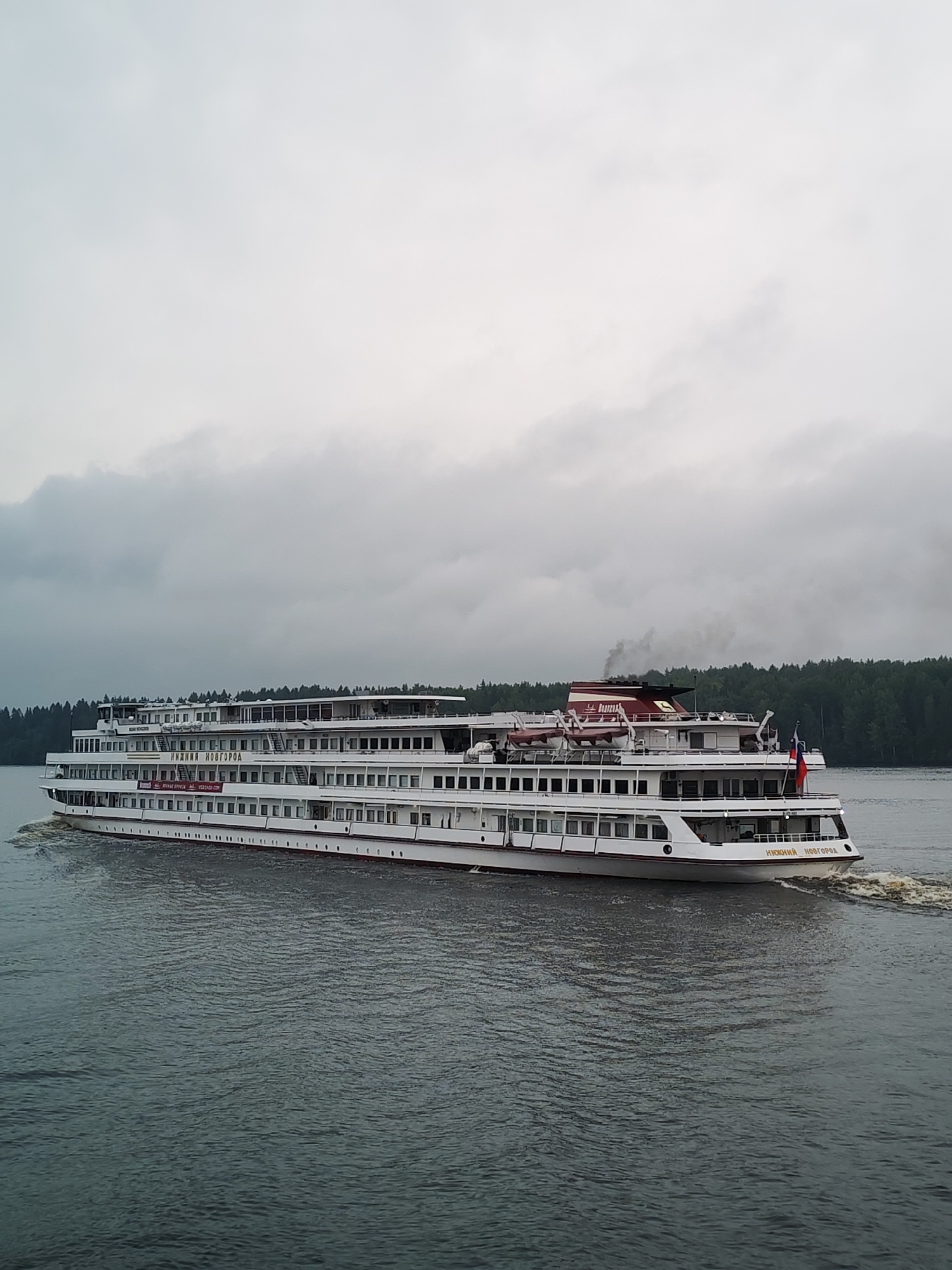 A large white passenger ship sails on a body of water under a cloudy sky. The ship has multiple decks with many windows. The name "НИЖНИЙ НОВГОРОД" is visible in gold lettering on the side of the ship. A red and white smokestack emits dark smoke. A Russian flag is visible on the stern of the ship. The water is dark and choppy, with a wake trailing behind the vessel. Dense green trees line the distant shore. The scene appears to be in the city of Khoroshevo, Russia. There are no people visible on the deck of the ship.