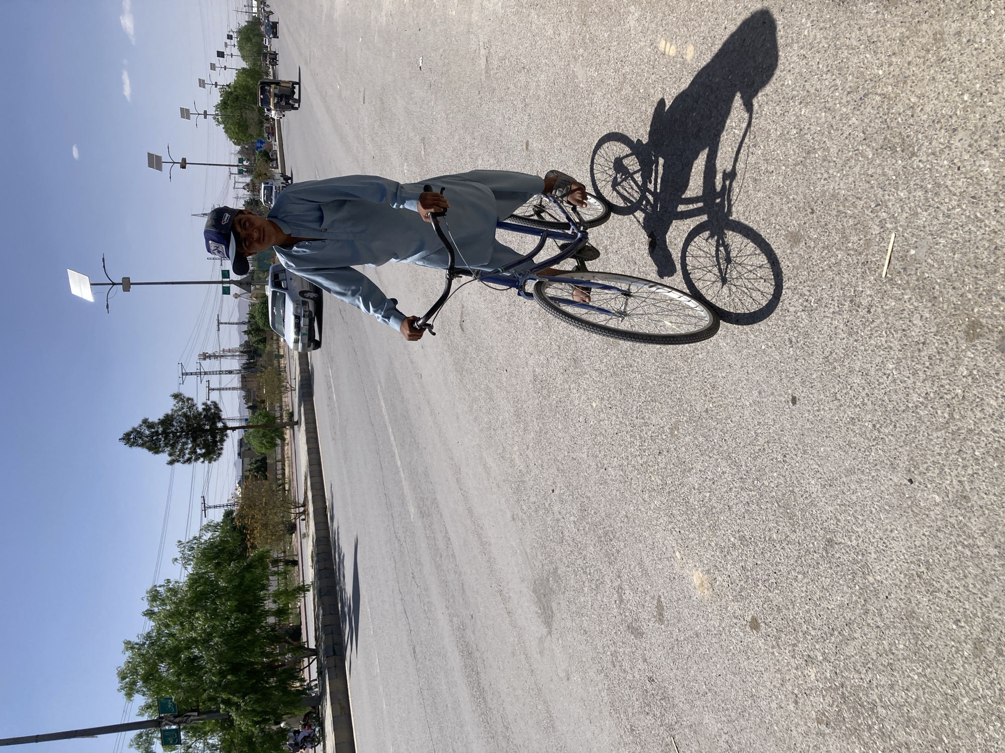 A young man, likely a teenager or young adult, is captured on a sunny day posing with a blue bicycle on a paved road. He is dressed in a light blue, traditional South Asian shalwar kameez and wears a blue and white baseball cap, facing the camera with a calm expression.
The strong, distinct shadows of the man and his bicycle stretch across the asphalt, indicating a high sun position, likely midday or early afternoon. The sky above is clear and bright with only a few faint clouds visible.
To the left of the road, a dirt shoulder gives way to a sidewalk lined with green trees of varying sizes. Utility poles, heavily laden with power lines, extend into the distance. In the background, other vehicles, including cars and motorcycles, are parked or moving further down the street, with distant buildings and hills completing the horizon. Modern solar-powered streetlights are also visible along the road. A small piece of debris lies on the asphalt near the bicycle's shadow. The scene depicts an outdoor setting consistent with an urban or semi-urban environment in Quetta City Tehsil, Pakistan. No visible text is discernible in the image.