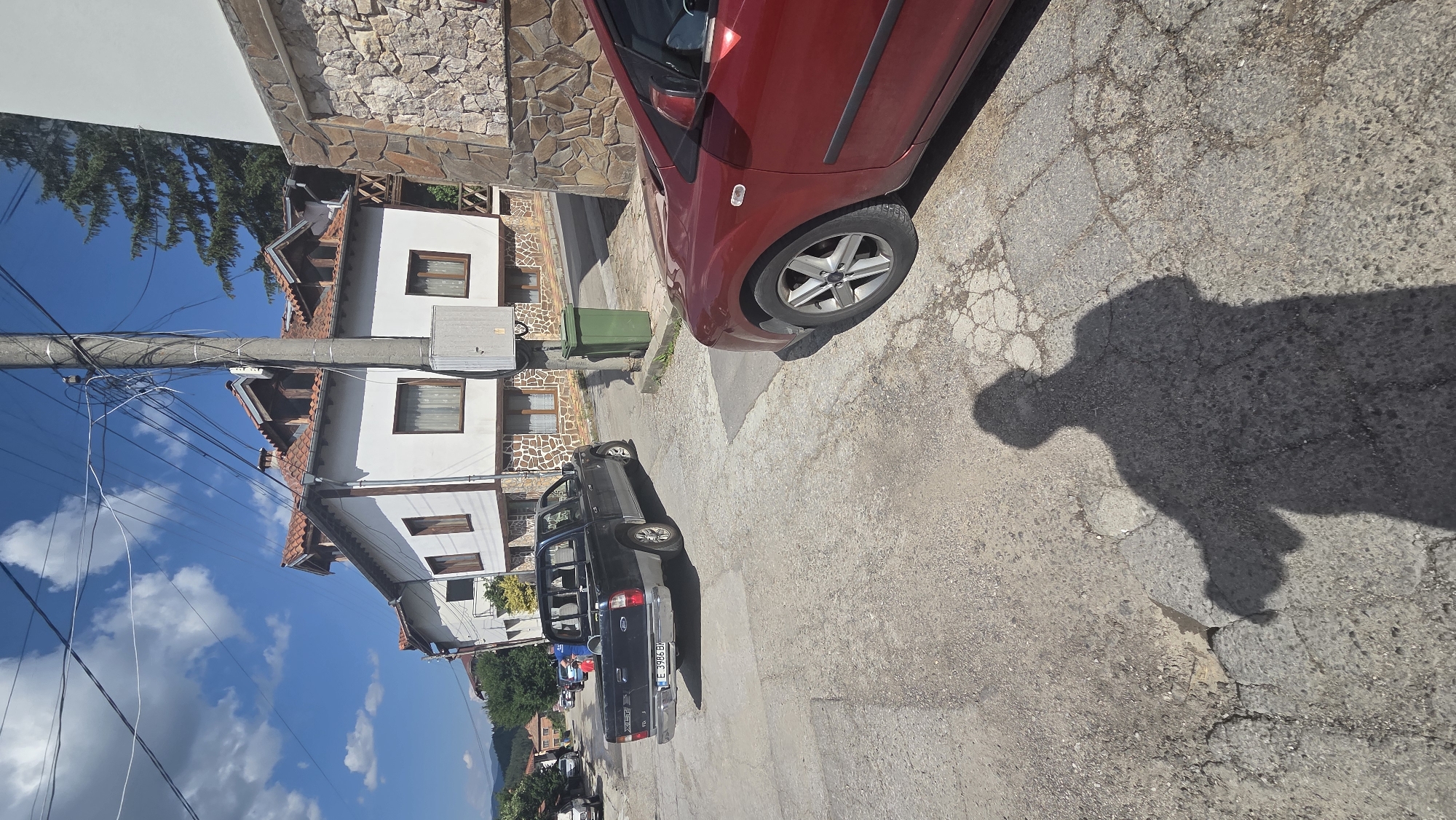 A shadow of a person is cast on a cracked asphalt road in an outdoor, urban environment. In the background, a black pickup truck is parked on the left side of the road, and a red car is parked on the right. Buildings with white walls and red tile roofs line the street. A utility pole with numerous wires stands near the center of the image, next to a green trash bin. The sky is blue with scattered white clouds. The scene appears to be a residential or commercial street in a town.