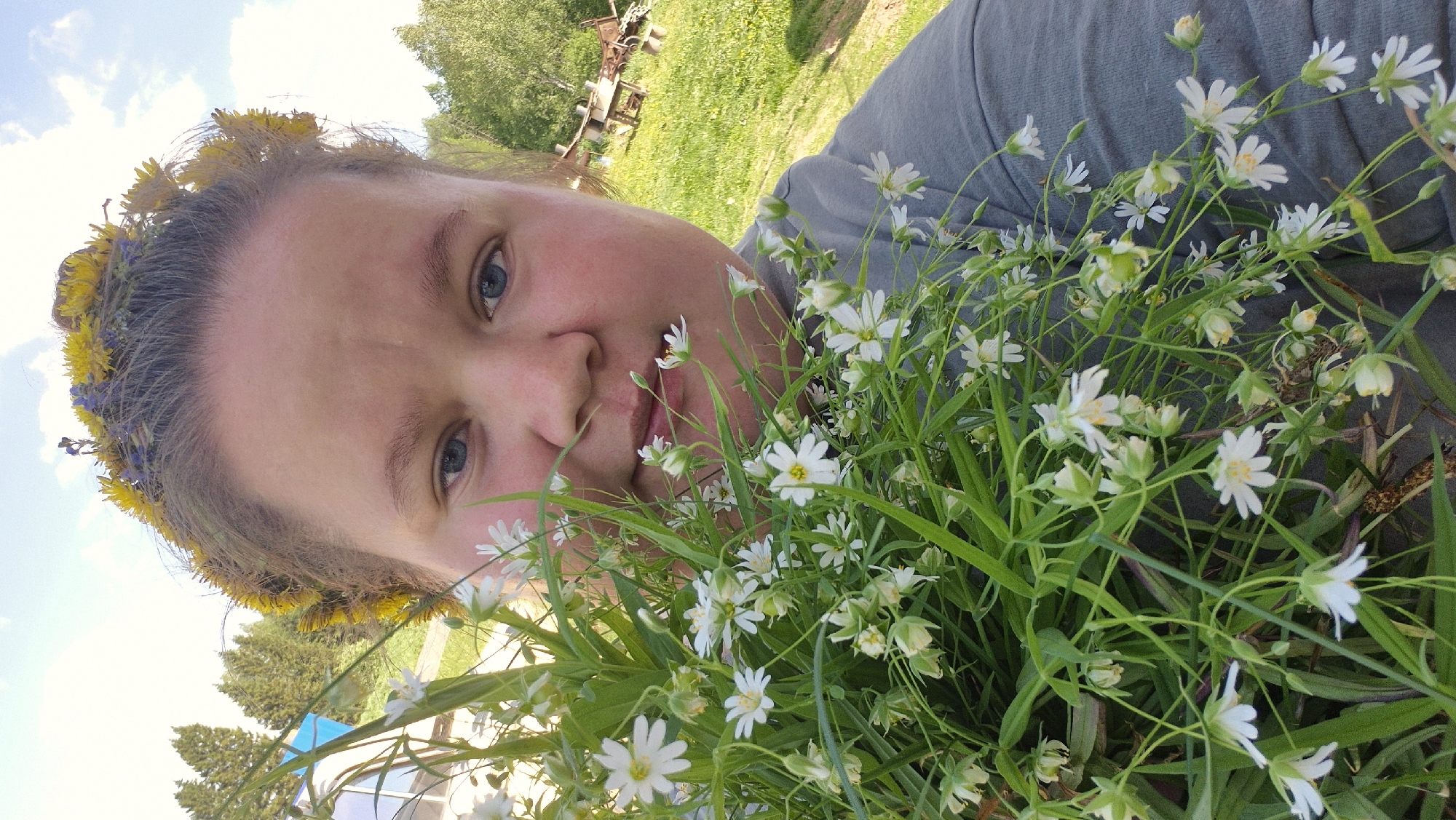 A medium shot depicts a person's face close to the camera, with a flower crown on their head and holding a bouquet of small white flowers. The person has fair skin, blue eyes, and is looking at the camera. The flower crown is made of yellow and blue flowers. The bouquet is composed of numerous small white flowers with green stems and leaves. The background includes grass, trees, and blue sky. The location is Дойная, Russia.