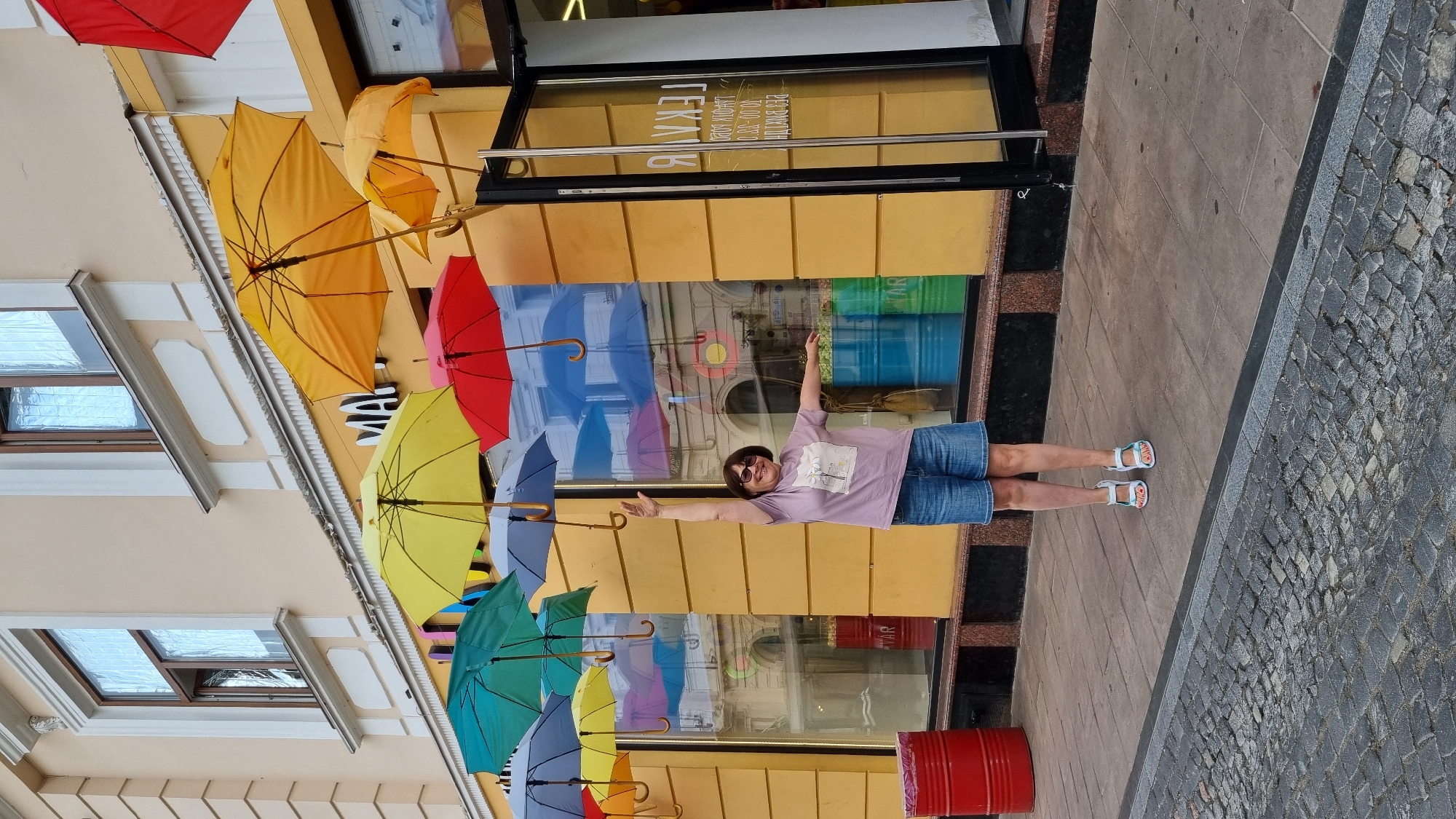 A woman stands on a sidewalk in front of a yellow building with large windows, her right arm raised in the air and her left arm extended to the side. She is wearing a light purple t-shirt, denim shorts, and white sandals. Above her, a colorful display of umbrellas hangs from the building's facade. The sidewalk is paved with square tiles and cobblestones. In the background, through the windows, shelves with products are visible, along with large cylindrical containers. Red and blue barrels are also present on the sidewalk. The image captures an outdoor urban scene, likely a street in Chernivtsi, Ukraine, given the context.