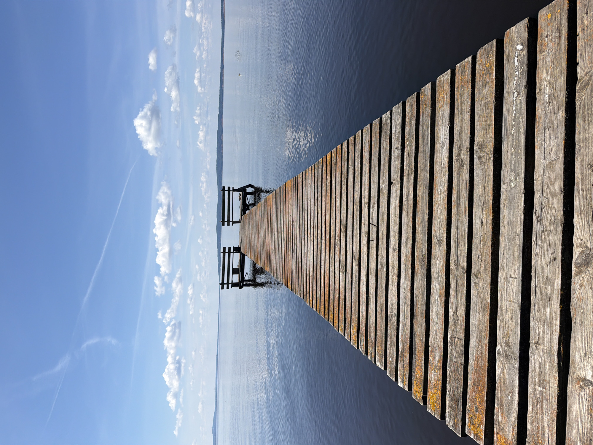 A wooden pier with benches at the end extends into a calm, dark body of water. The pier is made of weathered planks, some with orange discoloration. The sky above is a clear blue with scattered white clouds and a faint contrail. In the distance, a low, tree-covered shoreline is visible. The scene appears to be located in or near Stepanovo, Russia, likely a waterfront area on a sunny day. There are no people or animals visible in the image.