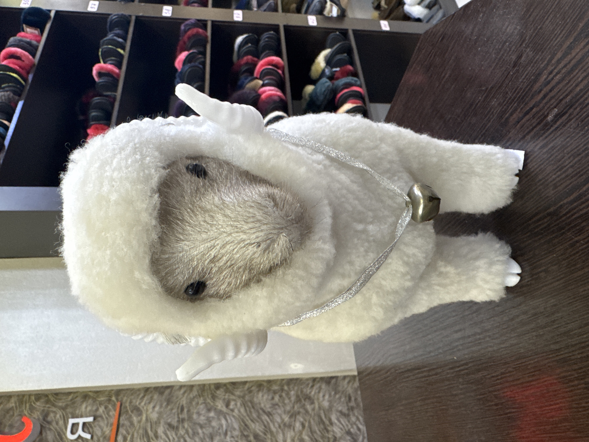 A stuffed sheep toy with white, fluffy wool and small, white wings stands on a dark wooden surface. A silver glittery ribbon is around its neck, with a golden heart-shaped bell attached. The sheep has black eyes and a brown, fuzzy face. In the background, shelves filled with various colored yarn balls are visible. To the left, a sign with Cyrillic letters can be seen. The setting appears to be a retail store or a craft shop.