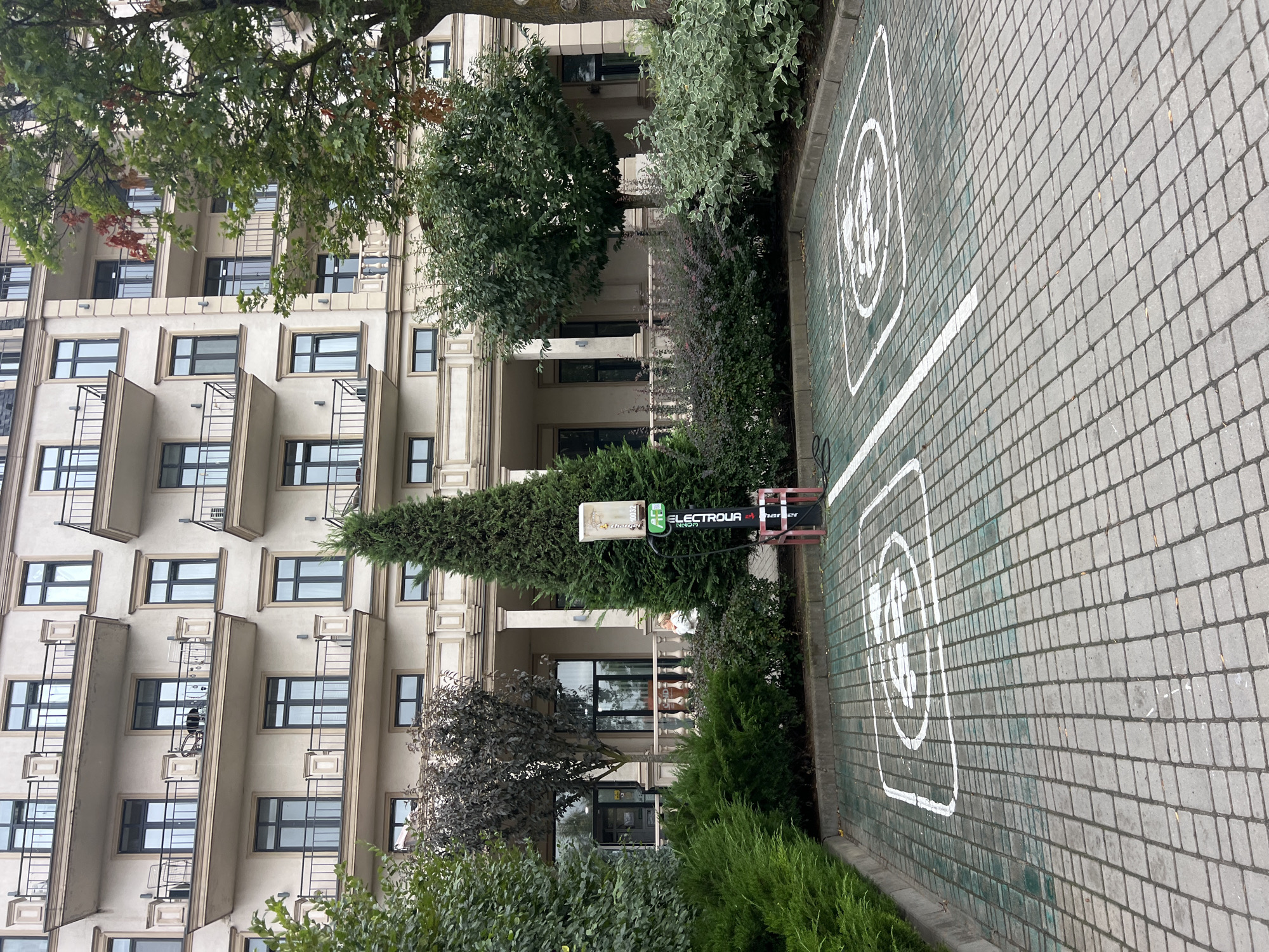 In Fontanka, Ukraine, an outdoor view captures a building facade with multiple balconies and windows, suggesting an apartment complex. In the foreground, a paved parking area features two marked spots, each with a white outline containing a symbol indicating an electric vehicle charging station. Between these spots, a charging station with the label "ELECTROUA" stands tall, connected to a power source. Lush green trees and bushes surround the parking area, creating a natural backdrop against the urban architecture.