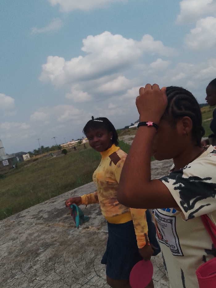 Three young women are walking in an open, grassy area in Umueshi, Nigeria, under a partly cloudy sky. One woman is in the foreground, wearing a beige T-shirt with a graphic print, a black bracelet with a pink charm, and carrying a red bag and a pink object. She has her hand on her head. Another woman in the background wears a yellow top with cut-out shoulders and a dark skirt, holding a blue and green cloth. The third woman is partially visible on the right. The environment includes a building and utility poles visible in the distance, and a concrete walkway or surface in the foreground.