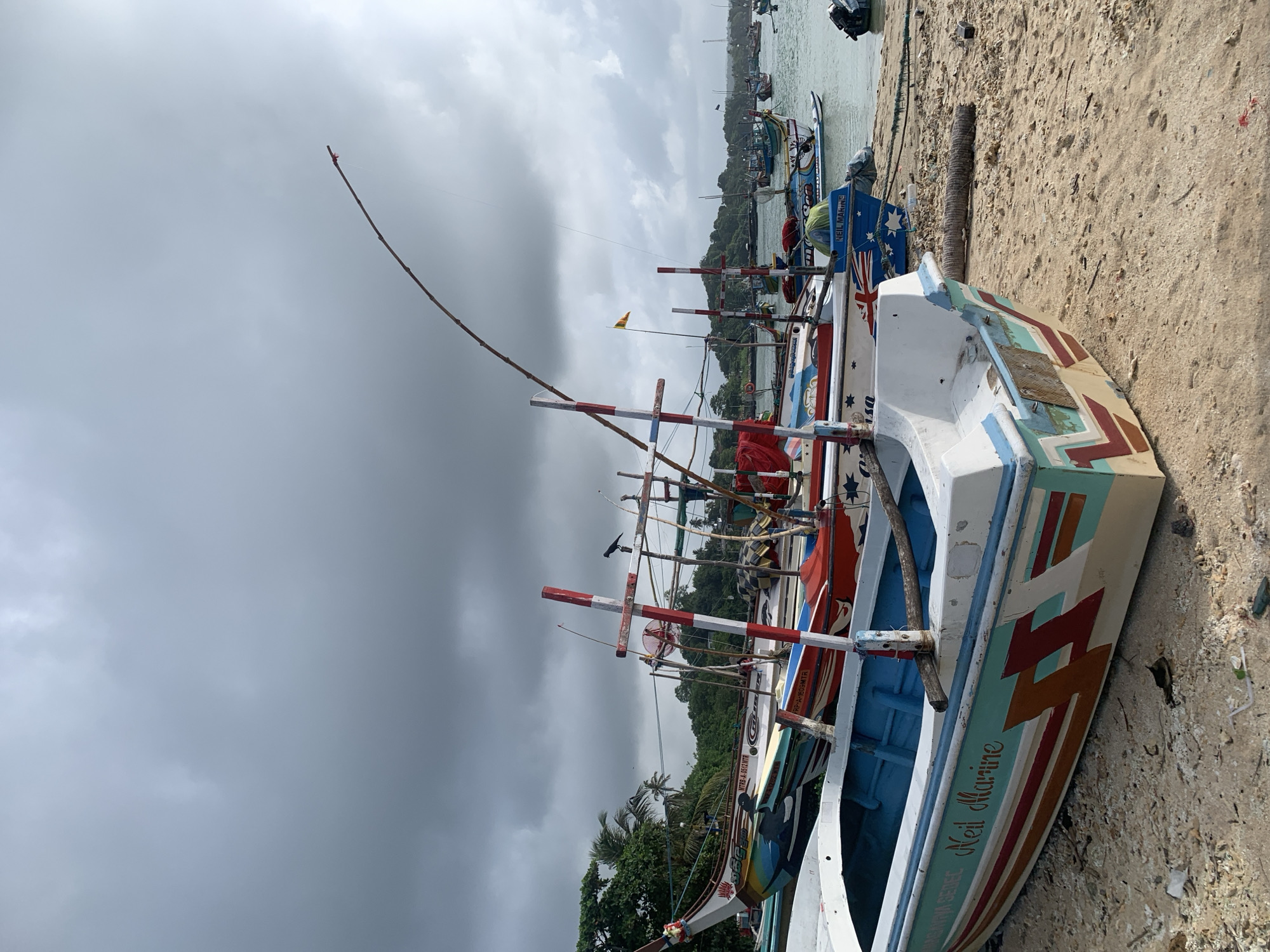The image depicts a harbor in Weligama, Sri Lanka, with numerous colorful fishing boats docked on a sandy beach and in the calm water. The sky is overcast, with dark clouds suggesting the possibility of rain. The foreground features several of these traditional fishing boats, some partially on the sand and others in the water. The boats are adorned with vibrant paintings and names, such as "Neil Marine" visible on one of the vessels. Fishing gear, including poles and nets, are present on some of the boats. In the background, a line of trees and lush vegetation can be seen along the coastline, with more boats scattered across the water, some further out towards the horizon. The overall scene captures the daily life and industry of a coastal fishing community.