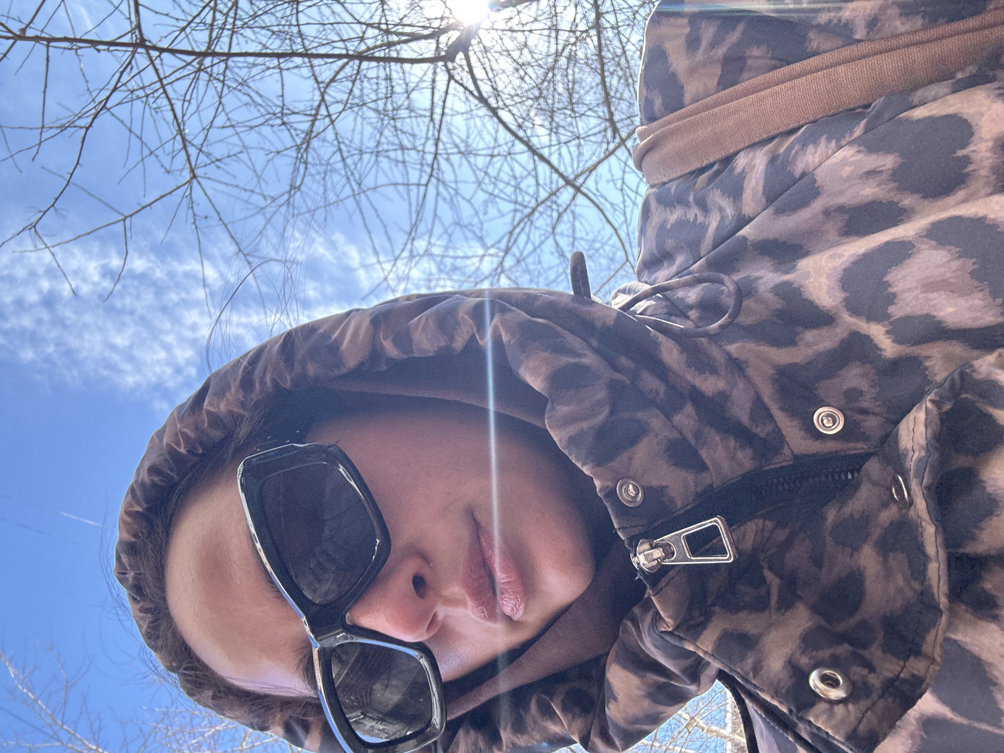 A low-angle selfie captures a person wearing sunglasses and a hooded, leopard-print jacket. The hood is up, partly obscuring their face. The sun shines brightly in the background, partly blocked by bare tree branches against a blue sky with scattered clouds. The zipper of the jacket is visible near the person's neck. The location cannot be determined from the given image.