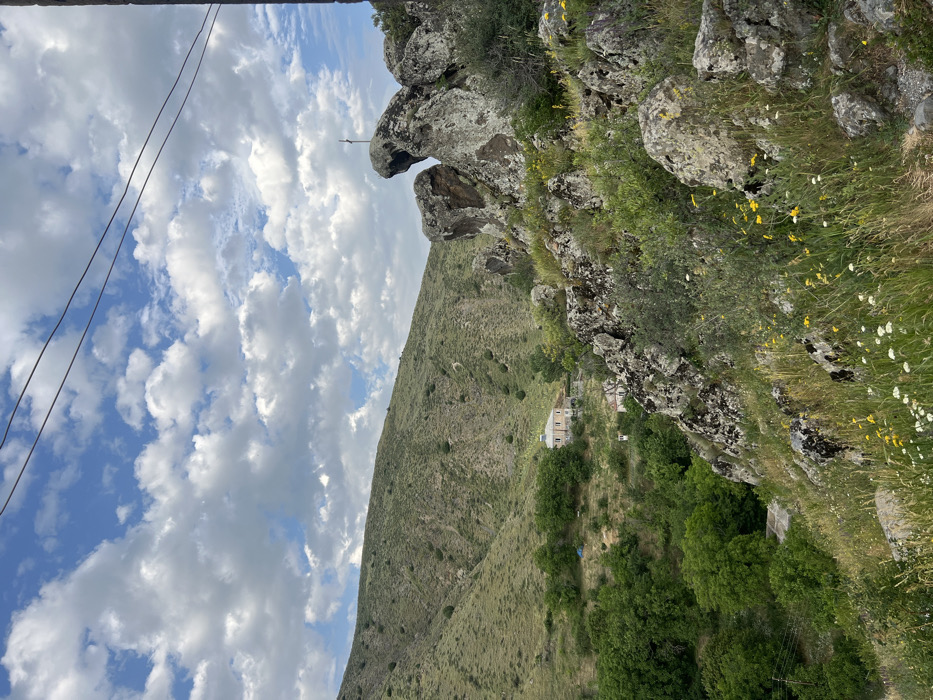 A mountainous landscape under a partly cloudy sky. The foreground features rocky terrain covered in vegetation. A hill slopes downward, dotted with trees, leading to a white building nestled among the greenery. In the upper left corner, power lines are visible against the sky. A unique rock formation resembling an animal head juts out from the top of the mountain.