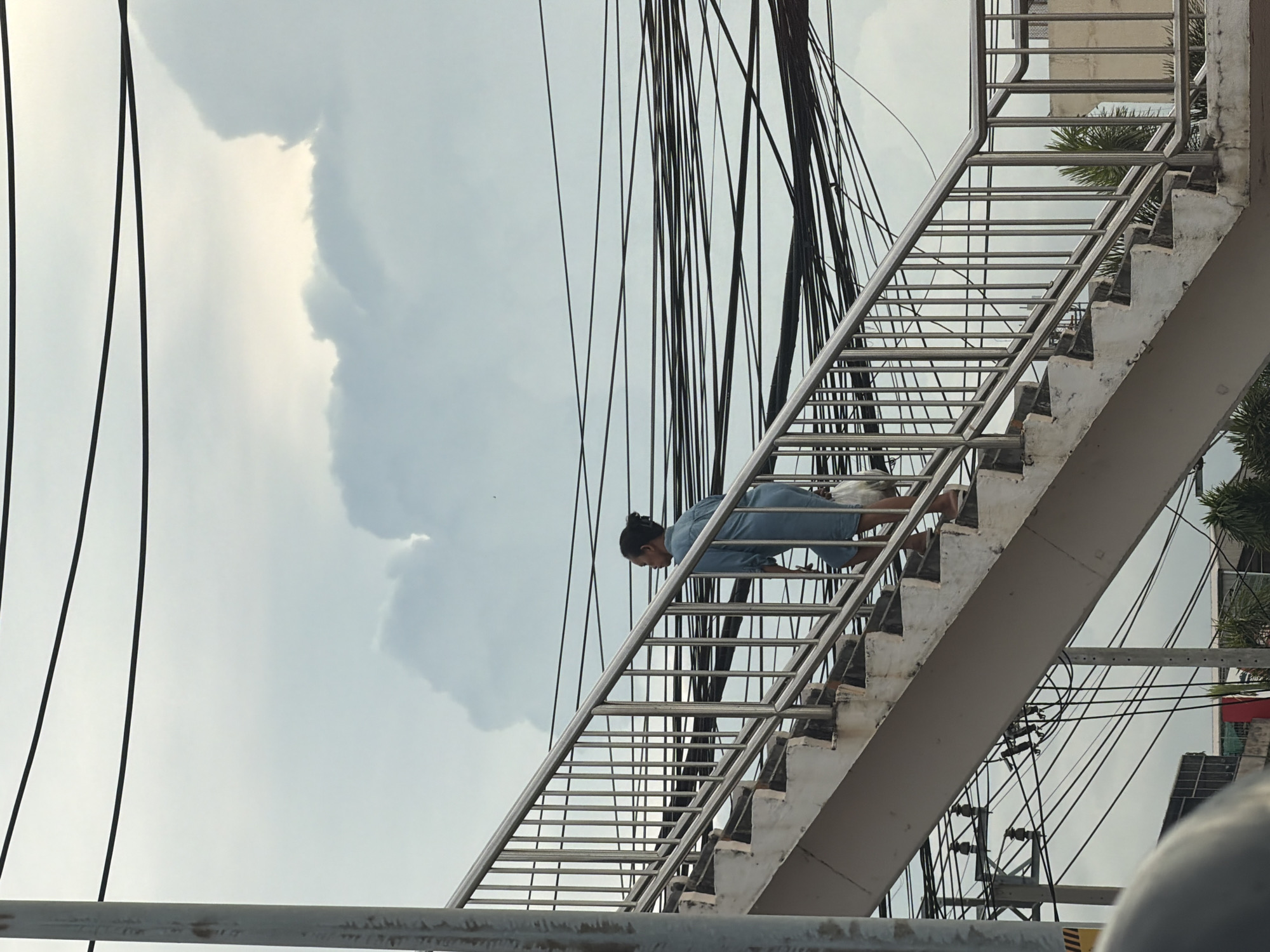 In an outdoor scene in Bangkok, Thailand, a woman with her hair in a bun, wearing a light blue shirt and denim shorts, ascends a concrete pedestrian overpass with a metal railing. She carries a translucent plastic bag in her right hand. The overpass stairs are worn, with visible concrete edges. Above and behind the overpass, a dense network of black electrical wires stretches across the sky, which is filled with a mix of light gray clouds and patches of bright sky. Buildings and palm trees are visible at the bottom of the image.